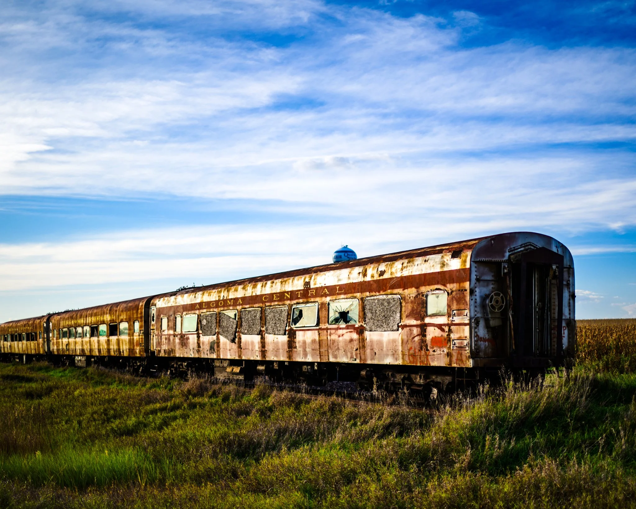 A row of abandoned, weathered passenger cars from the Algoma Central Railway rests beneath a wide blue sky, their rusted surfaces and fading paint suggesting years of quiet neglect.