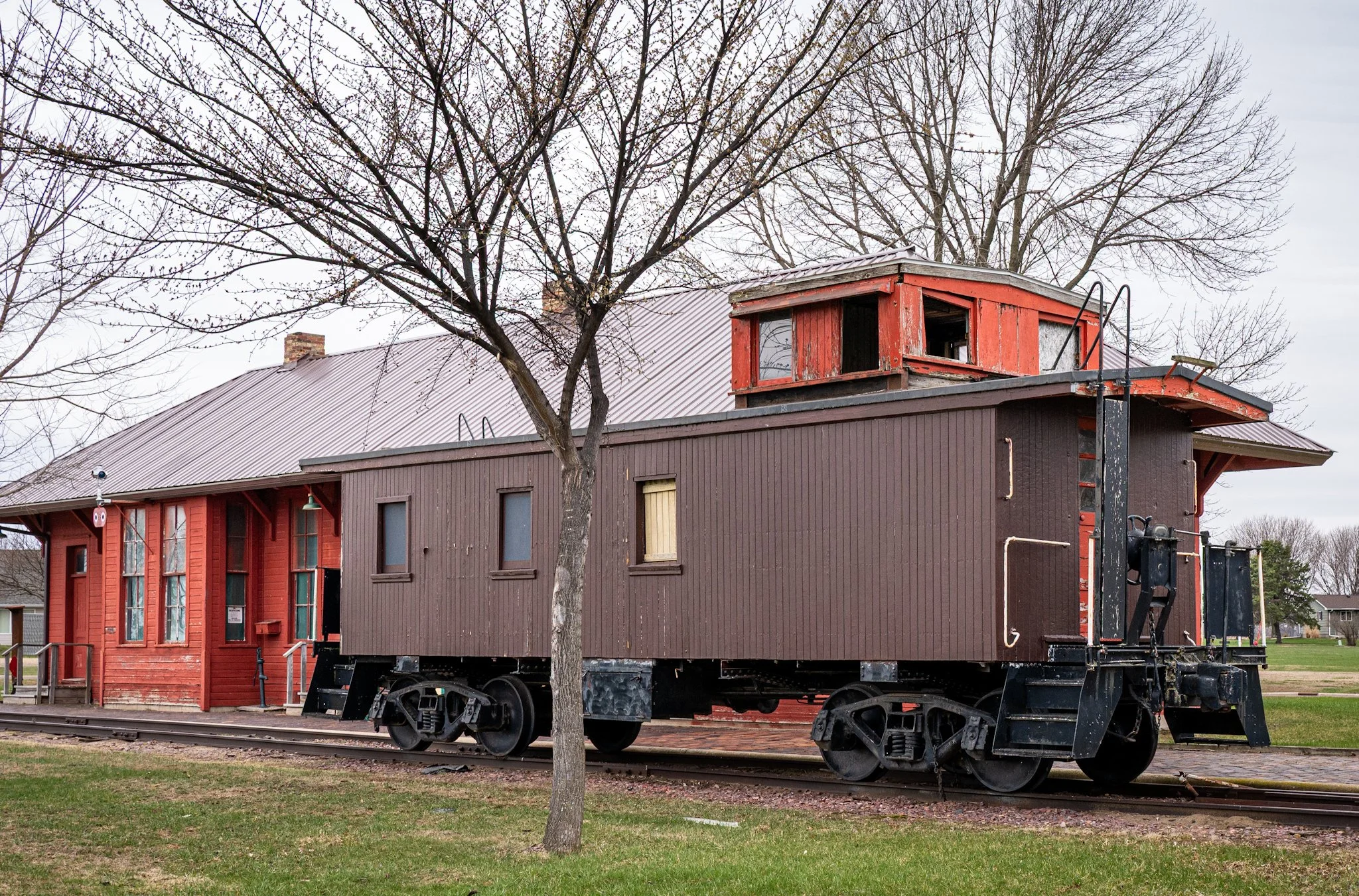 A red caboose sits on display near bare trees and a small red building, its preserved exterior evoking the history and nostalgia of older rail travel.