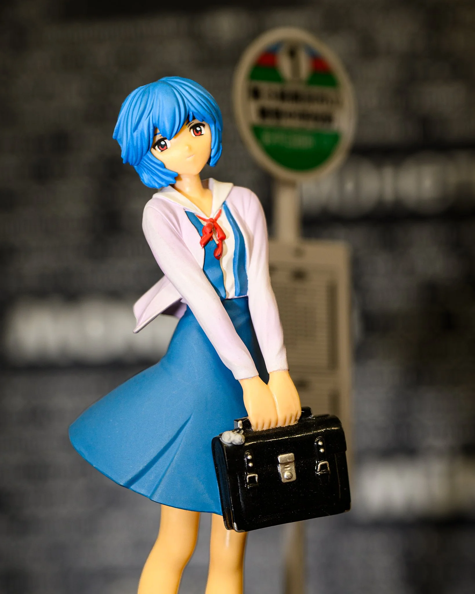 Anime figure of Rei Ayanami from Neon Genesis Evangelion standing with a slight head tilt and holding a black school satchel. She wears her white-and-blue school uniform, framed by softly blurred station scenery with a vintage railway sign in the bac