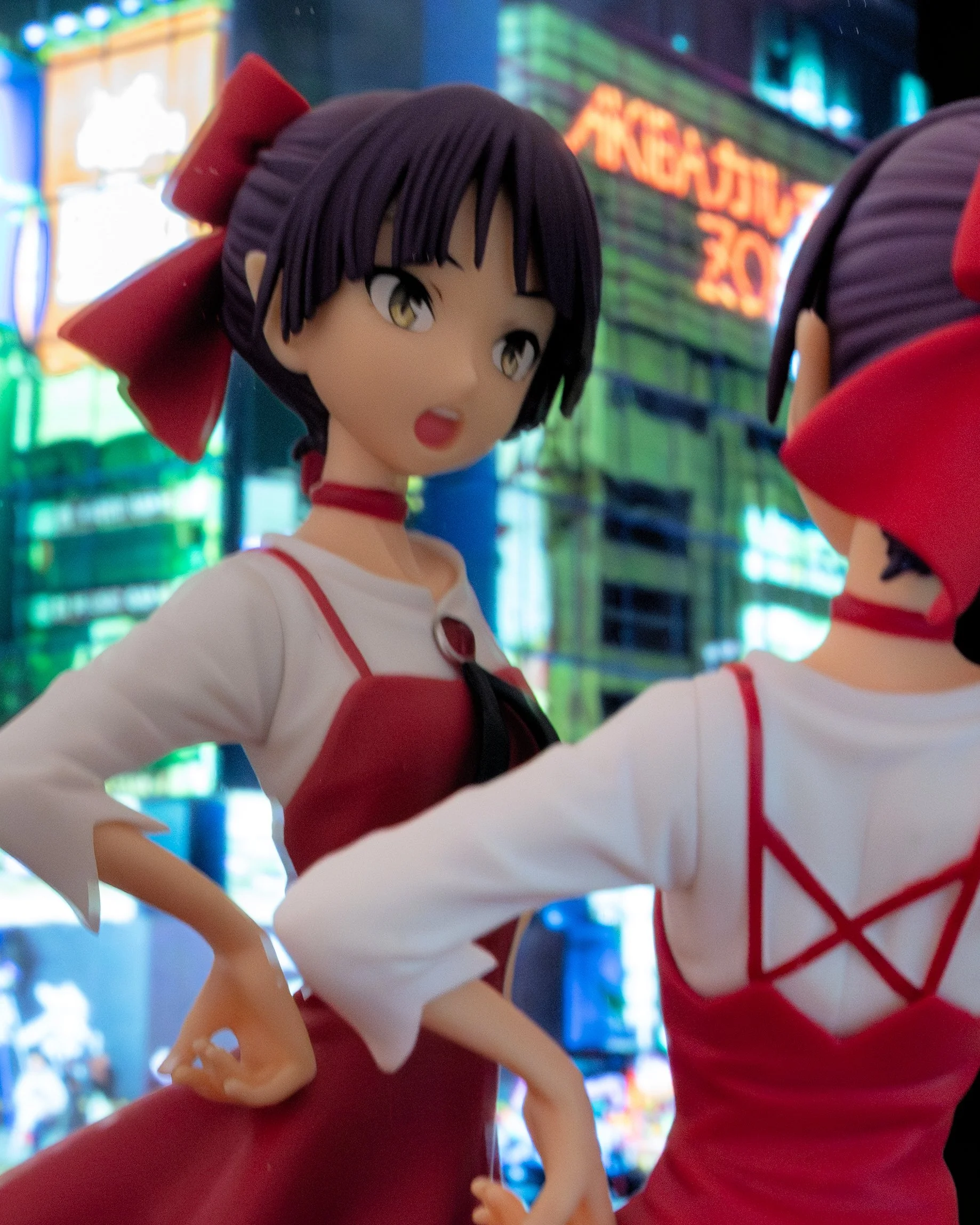 A stylized anime figure of a girl in a red outfit is posed in front of a mirror, showing both her front and back. Behind her, a blurred neon cityscape displayed on a screen creates a colorful nighttime backdrop. The scene has a soft, slightly dreamy 