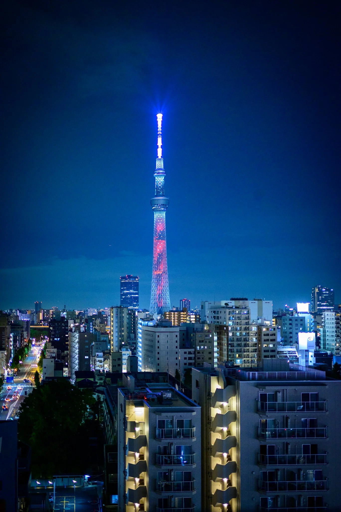 Night view of Tokyo Skytree rising above a dense cityscape, illuminated in blue and red. Surrounding mid-rise apartment buildings glow with warm interior lights, while streets below form lines of headlights and neon, all set beneath a deep blue night