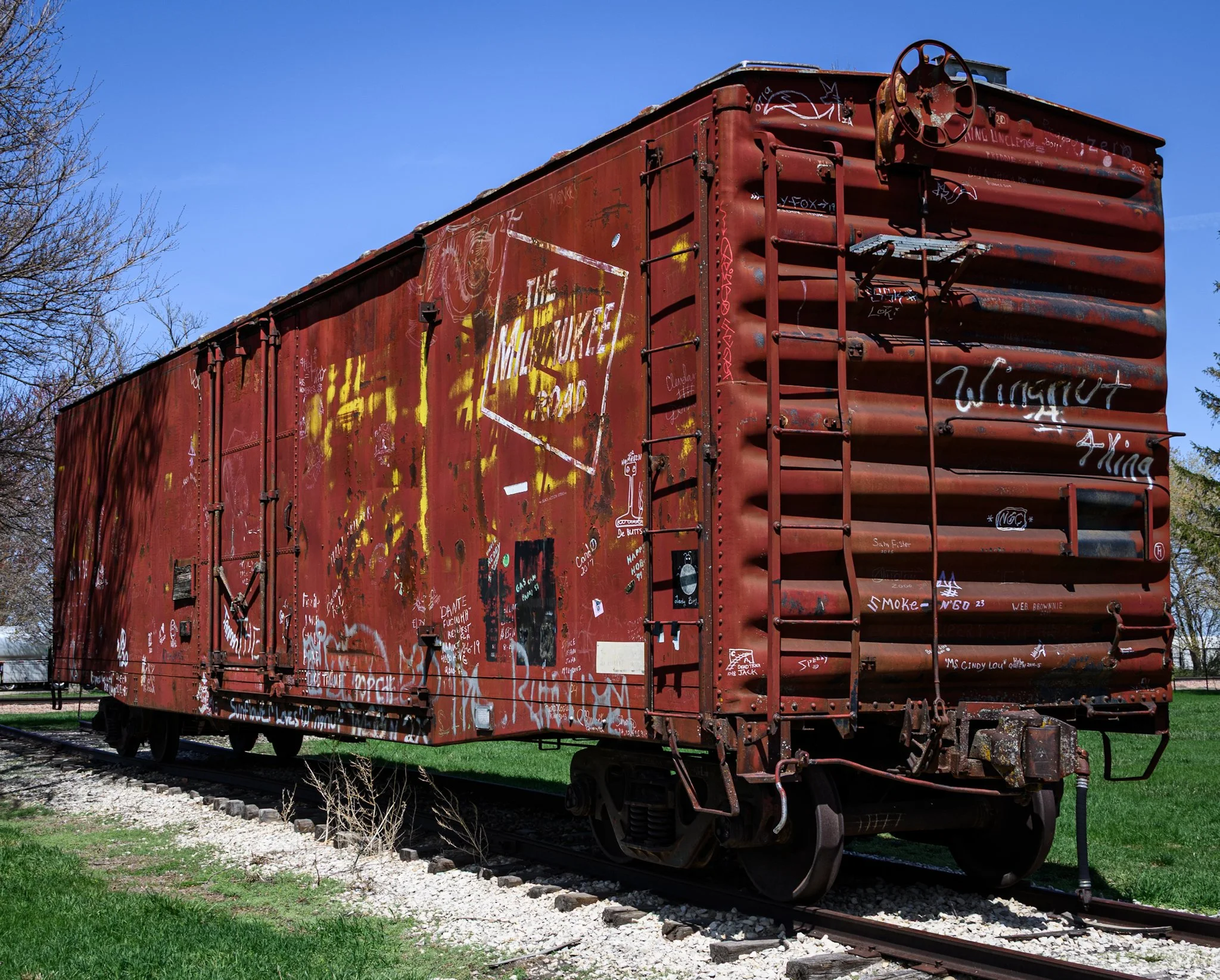 A weathered red Milwaukee Road boxcar sits on short display tracks in Britt, Iowa, under a deep blue spring sky. Rust, faded lettering, and layers of graffiti cover the historic railcar, which serves as a stage during the town’s Hobo Days festival an