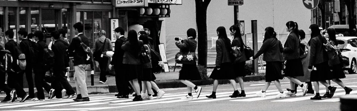 Black-and-white street photograph of students and commuters crossing a wide zebra crossing in Tokyo. A line of schoolgirls in dark uniforms moves from right to left while other pedestrians gather on the opposite side near a FamilyMart convenience sto