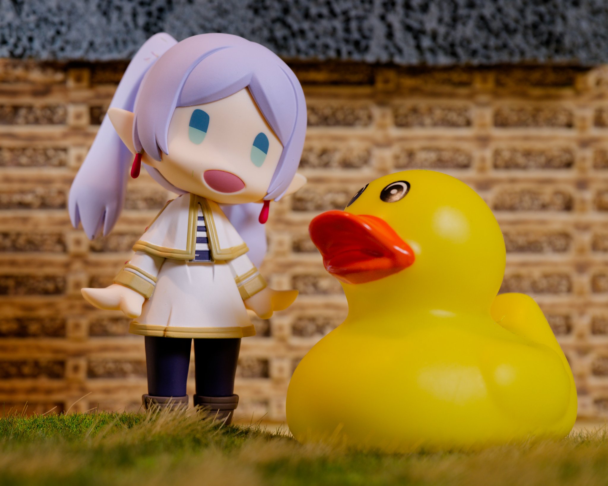A figure photograph of Frieren from Frieren: Beyond Journey’s End standing beside an oversized yellow rubber duck, both posed on a miniature outdoor scene. Frieren tilts her head toward the duck, while a wooden retaining wall and grass made from HO-s
