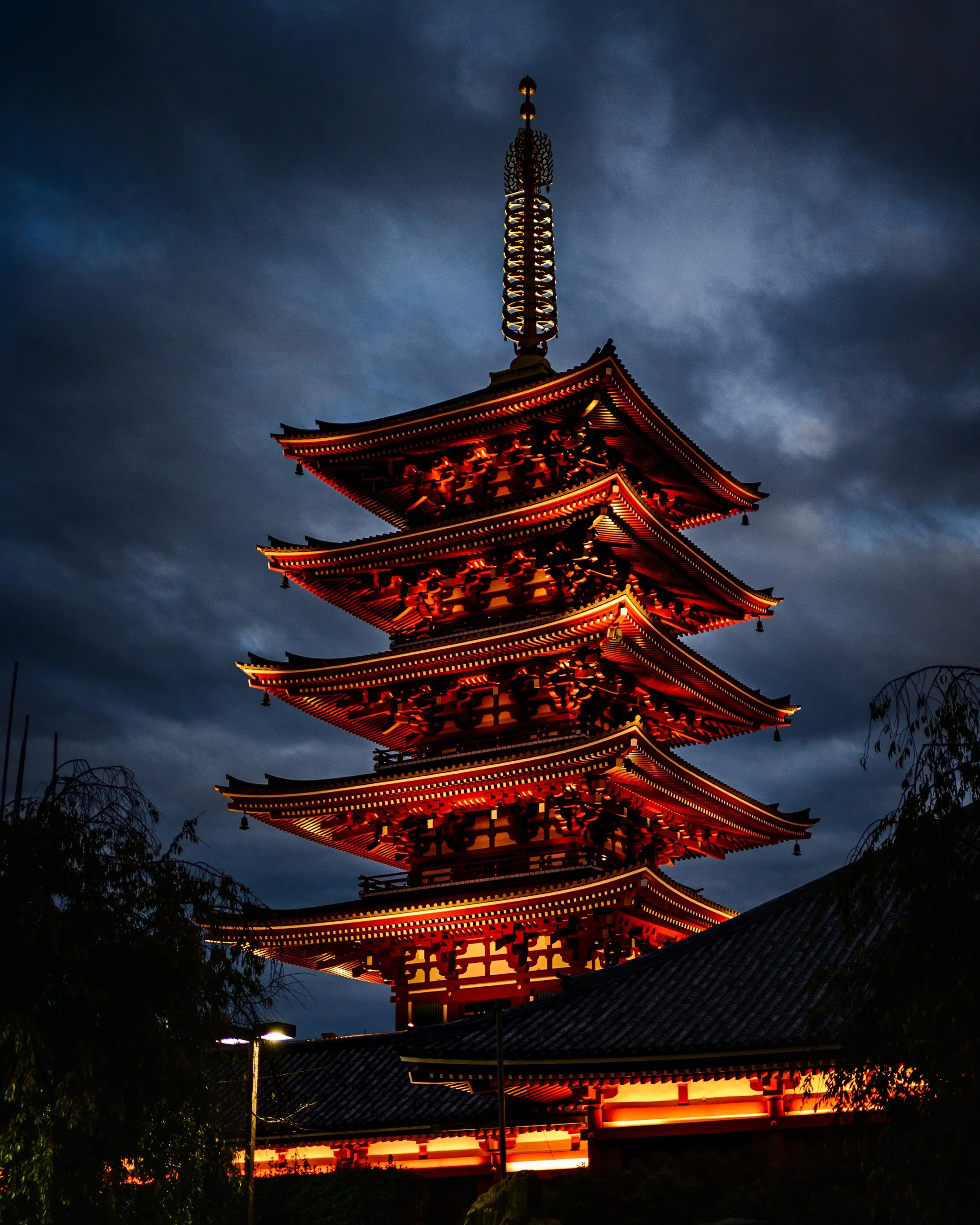A five-story pagoda illuminated in warm orange light rises against a dark, cloud-filled evening sky. The glowing tiers contrast with the cool blue-gray clouds, while silhouetted trees and traditional rooftops frame the lower portion of the scene.
