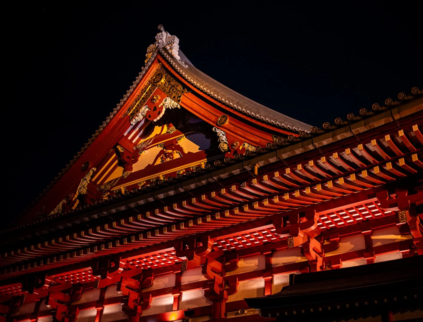 A detailed nighttime view of a traditional Japanese temple roof, glowing in rich red and gold tones against a deep blue sky. The image captures layered eaves, intricate wooden brackets, and ornamental carvings from a low angle, emphasizing the struct