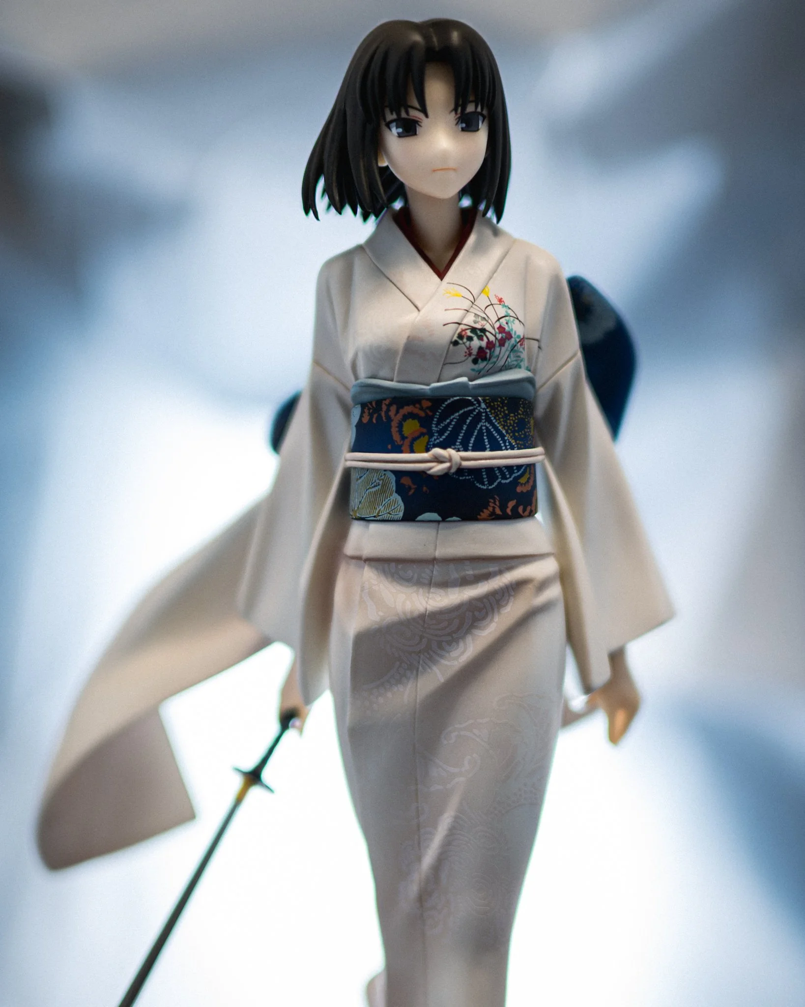 A detailed anime figure of Shiki from The Garden of Sinners stands in a softly glowing corridor-like space, wearing a pale patterned kimono with a dark blue floral obi and holding a sword at her side. The background is heavily blurred with cool blue-
