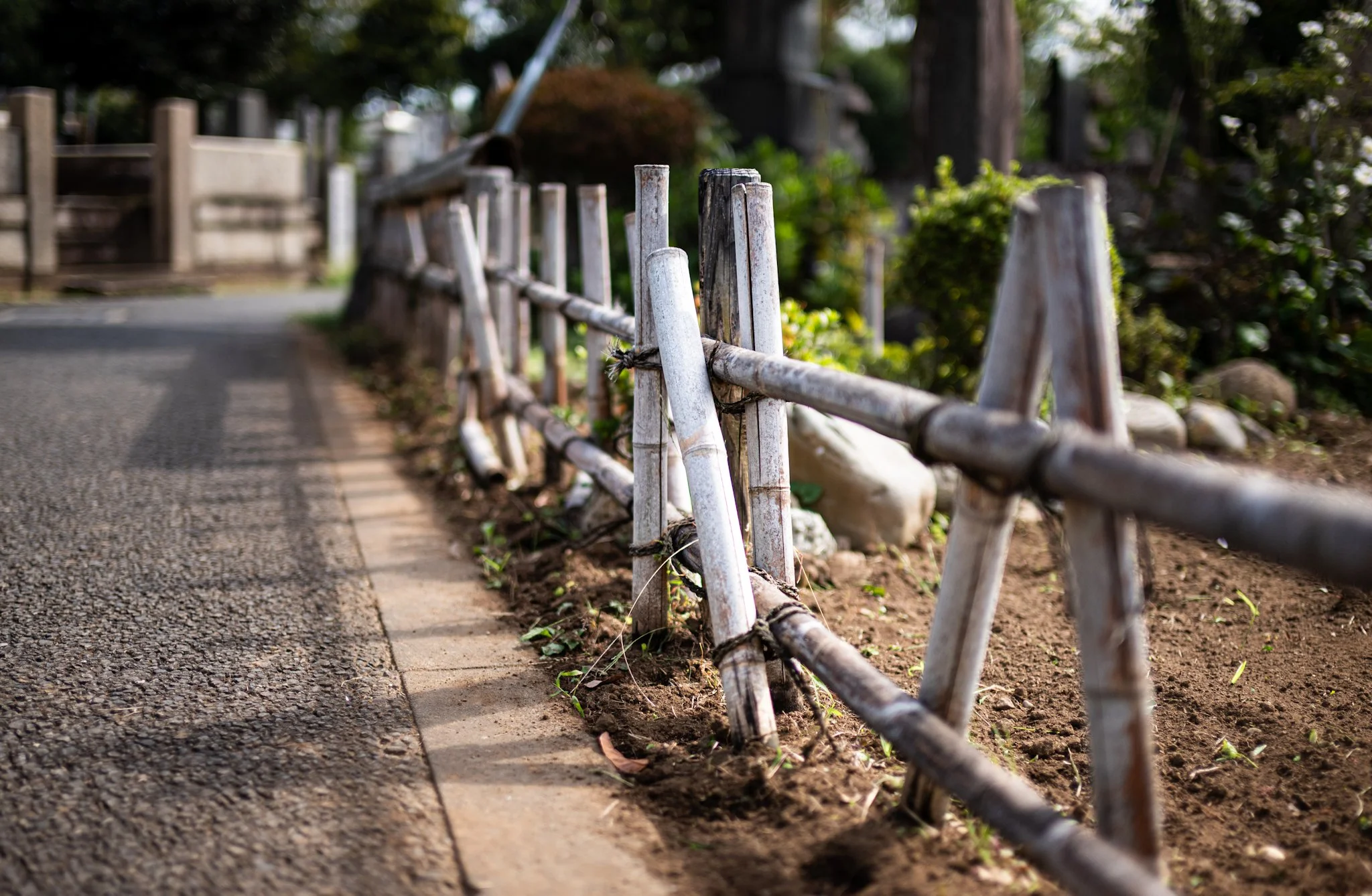 Shallow-depth-of-field view of a low wooden fence lining a narrow path, with weathered posts and rope ties in sharp focus while the walkway and surrounding greenery blur into the background in soft daylight.