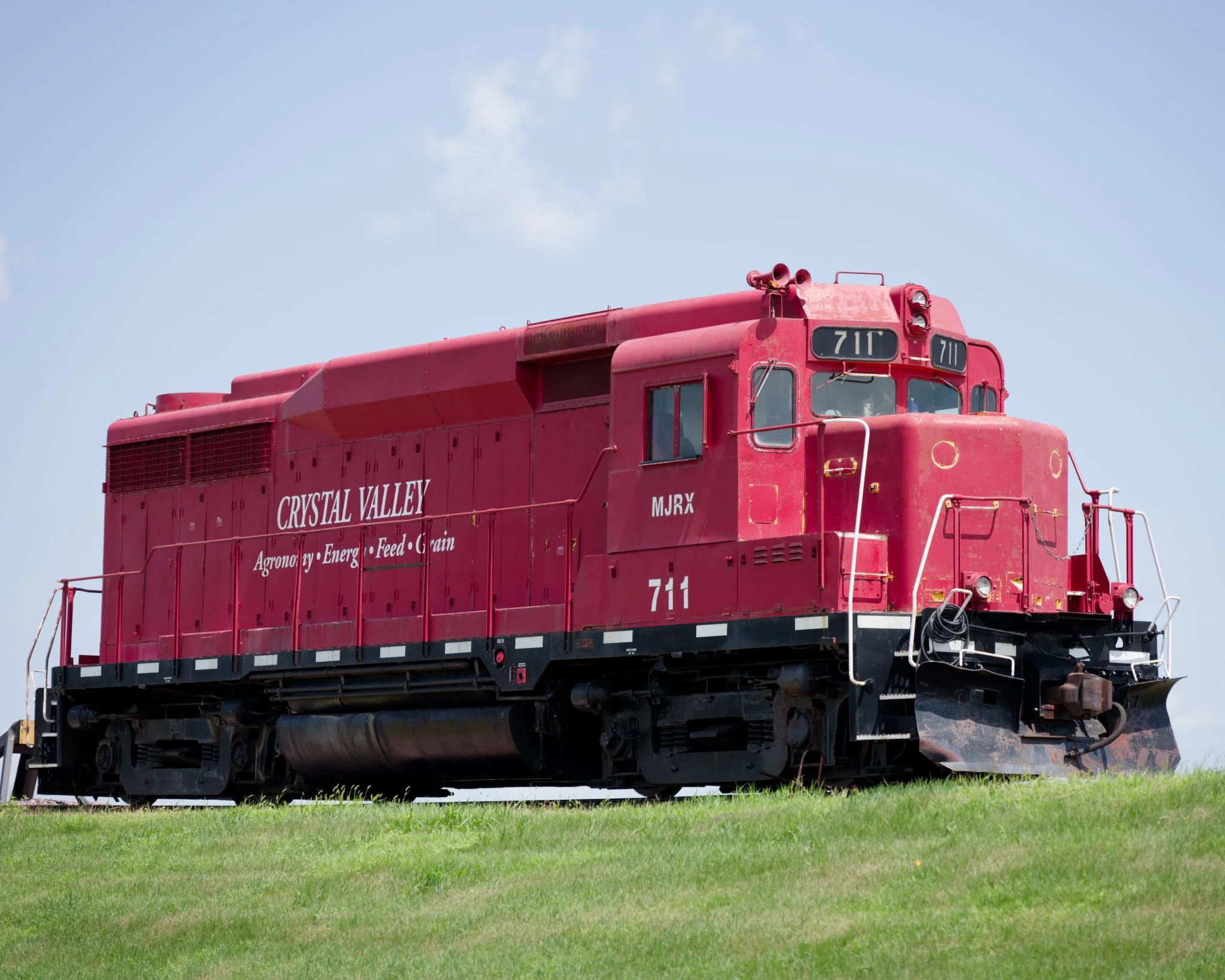 Low-angle view of red Crystal Valley locomotive 711 resting on a railroad spur, photographed from a mowed grassy ditch. The weathered locomotive rises against a pale blue sky, with white handrails, lettering, and mechanical details visible along its 