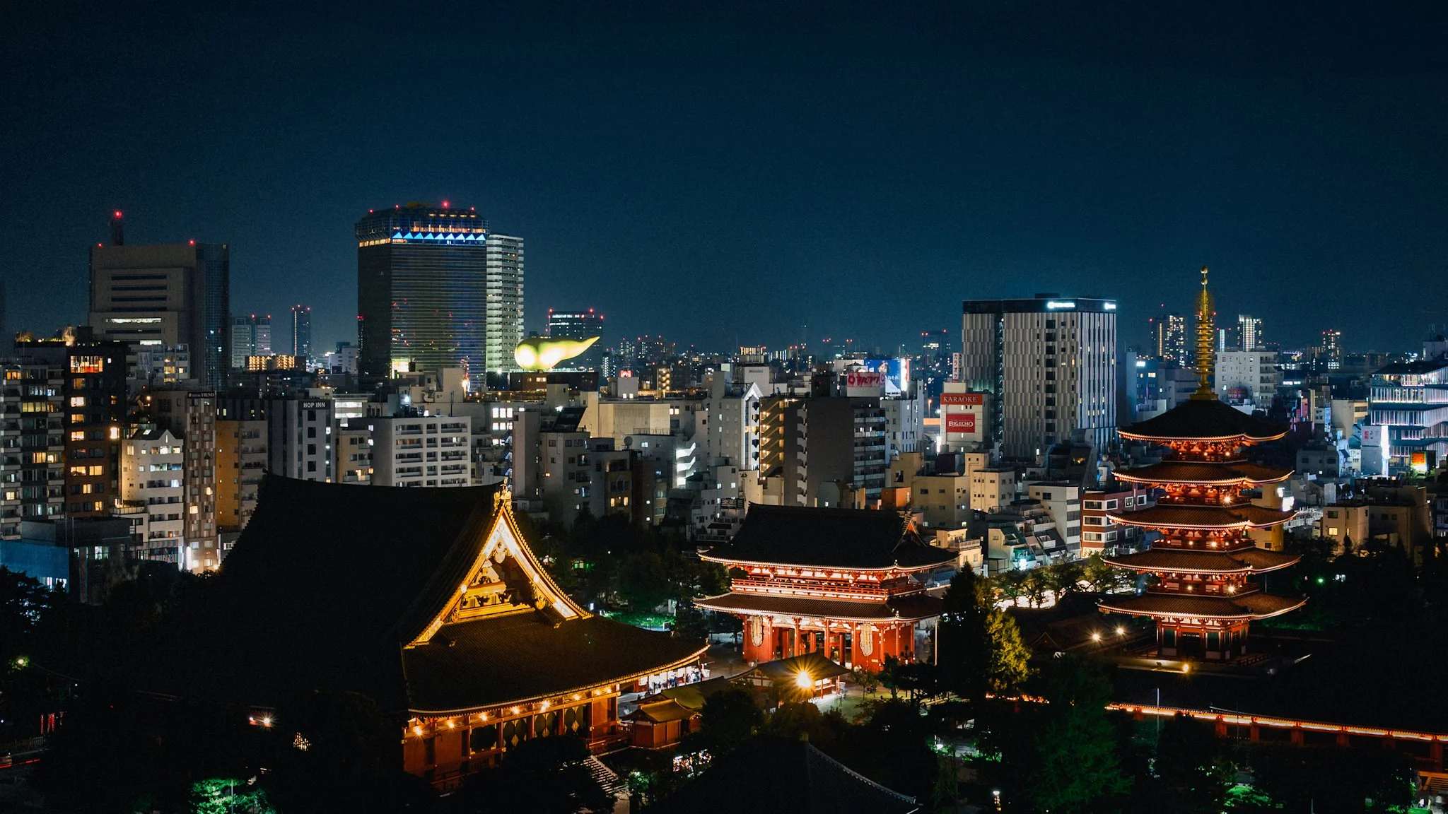 Night view of Sensō-ji in Tokyo, with the temple’s main hall and five-story pagoda glowing in warm orange light. Traditional rooftops and gates stand in contrast to a dense backdrop of modern city buildings and neon-lit signs, all set beneath a deep 
