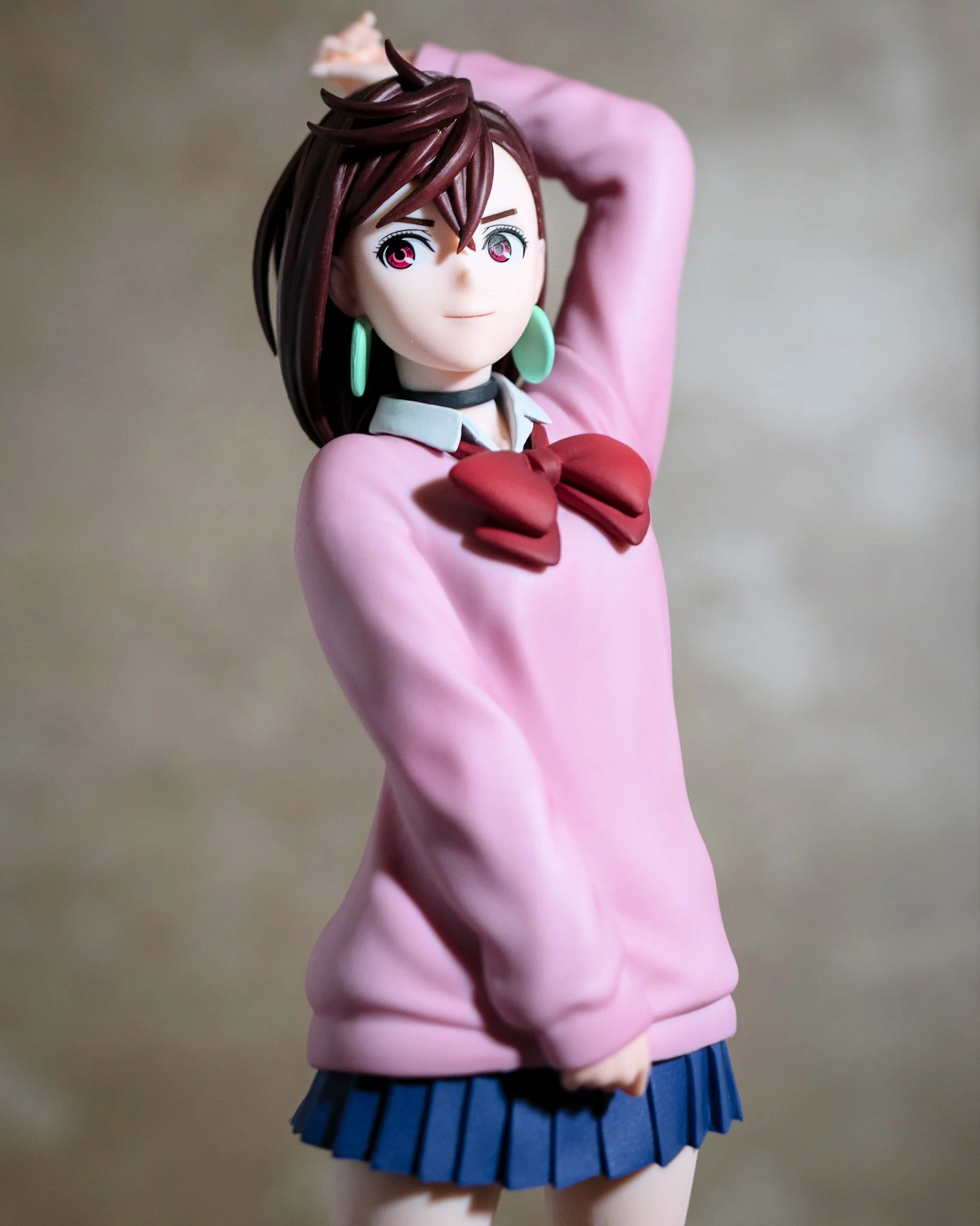 A stylized anime figure of a girl with short brown hair and teal earrings, wearing a pink sweater with a red bow and a blue skirt. She poses with one arm raised behind her head, softly lit against a smooth gray background, with the lighting emphasizi