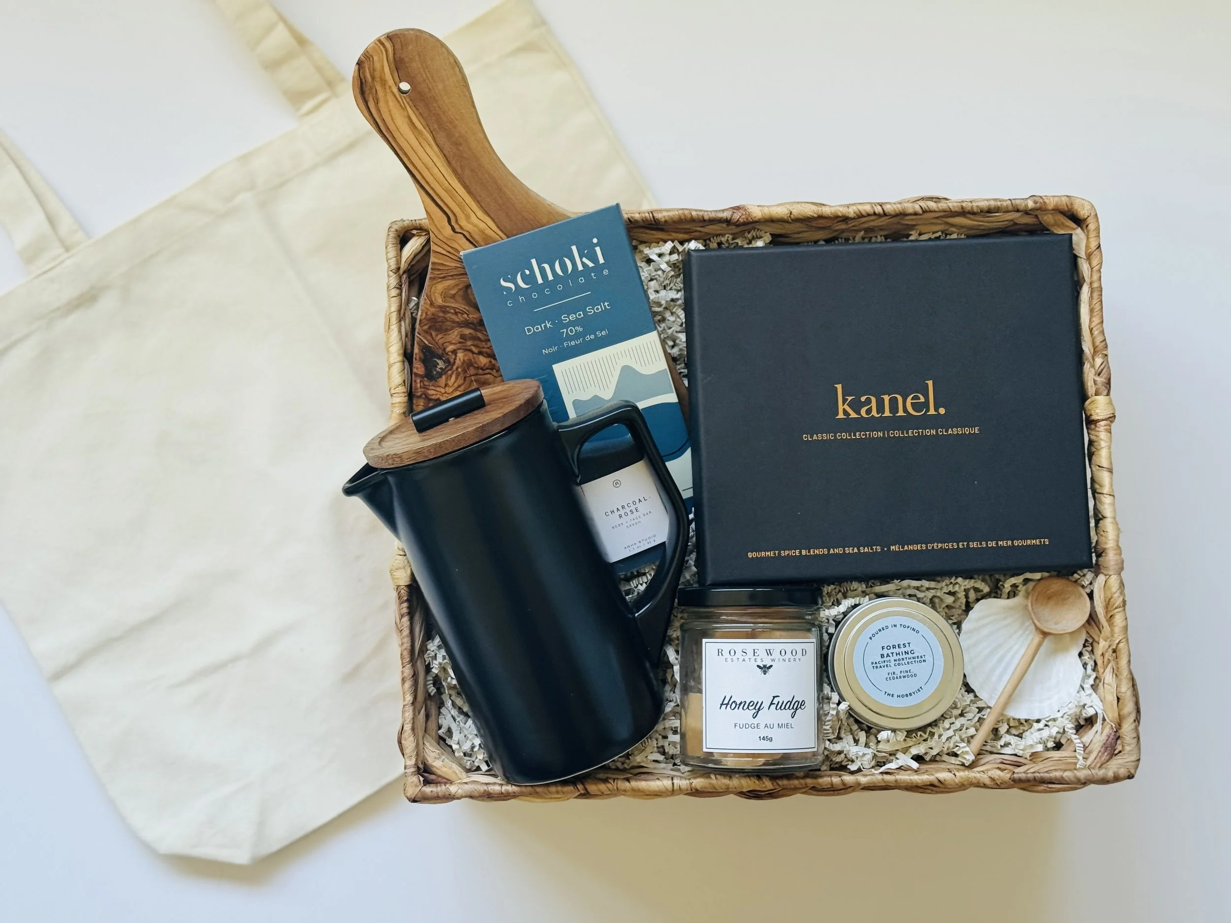 A gift basket containing a black teapot with a wooden handle and lid, a box of Kanel spice blends, a bar of Rosewood Honey Fudge, a tin of Forest Bathing balm, a blue Selvage chocolate bar, and a white seashell with a small wooden scoop, all arranged on shredded paper inside a woven basket.
