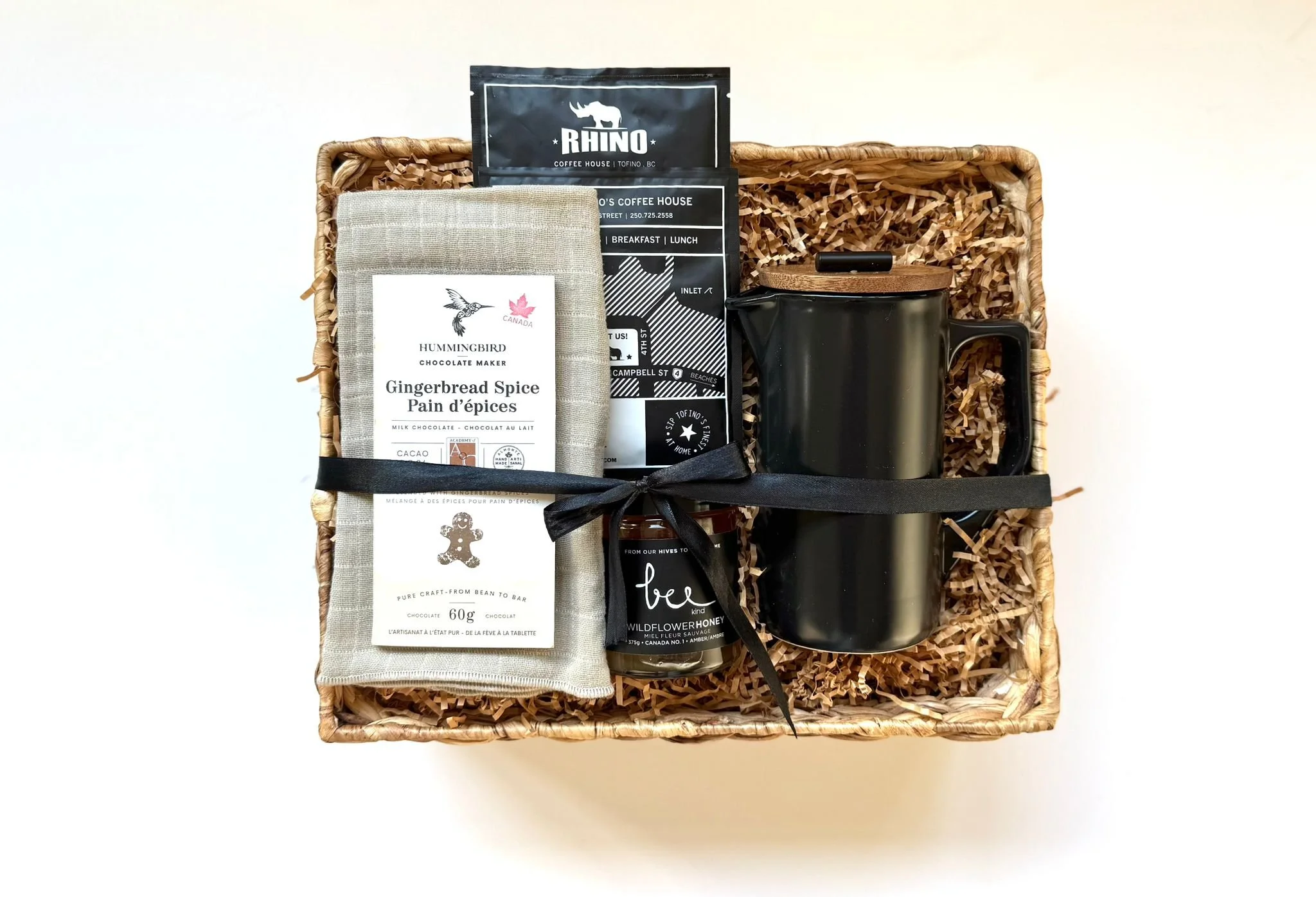 A gift basket containing a black mug with a wooden lid, a packet of gingerbread spice, a black milk chocolate bar, and a prayer candle in black with white text, all arranged on shredded paper inside a woven basket.