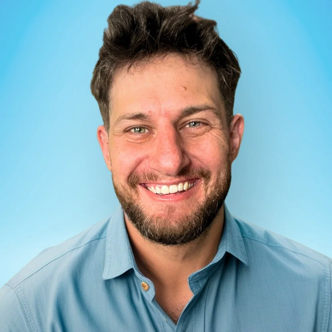 A smiling man with short brown hair and a beard, wearing a light blue button-up shirt, posing against a light blue background.