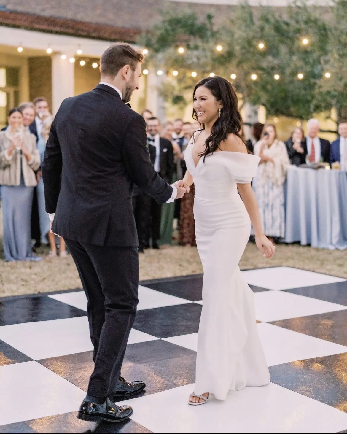 A couple dancing on an outdoor checkered floor, surrounded by guests, with string lights overhead.