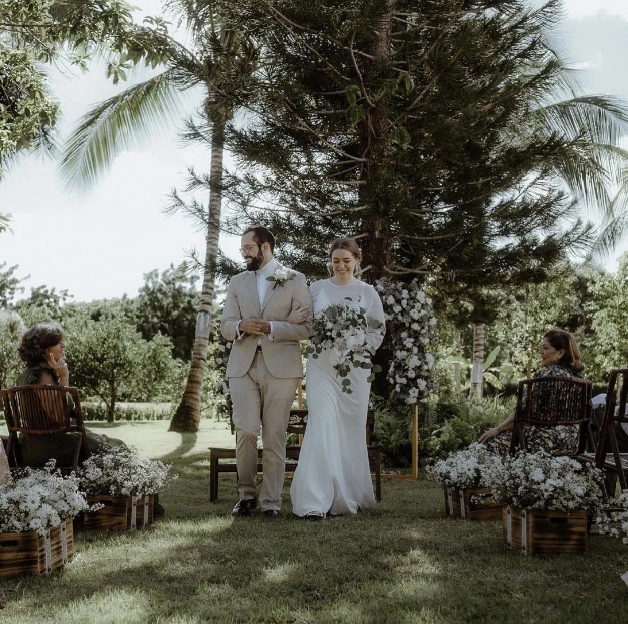 Bride and groom walking down an outdoor aisle surrounded by trees and chairs, with guests seated and floral decorations.