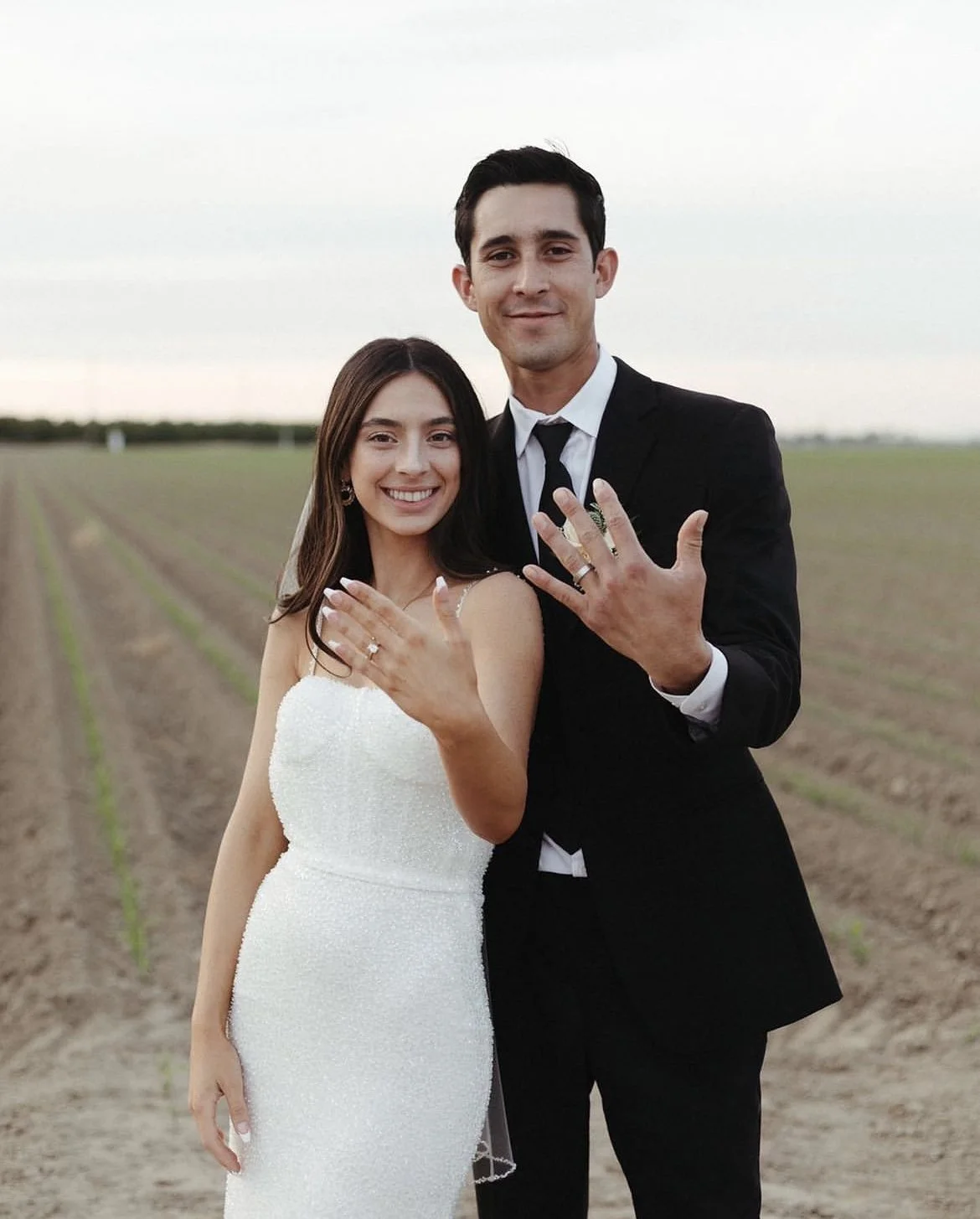 A couple in wedding attire standing in a field showing their rings
