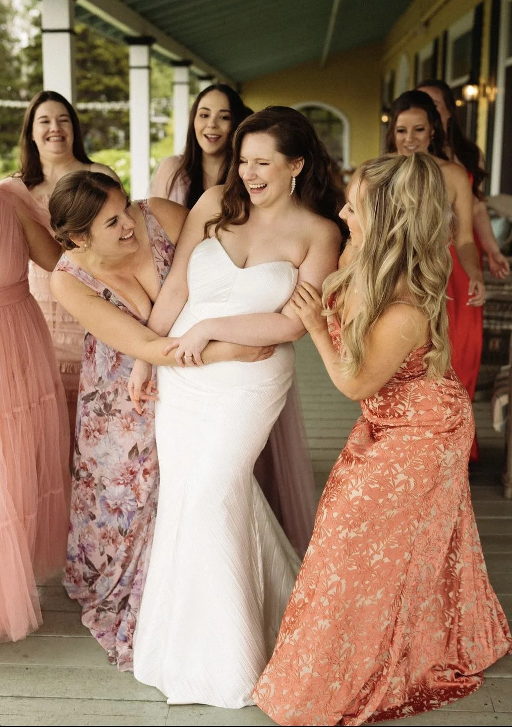 A bride in a white dress surrounded by bridesmaids laughing and interacting, wearing floral and colorful dresses on a porch.