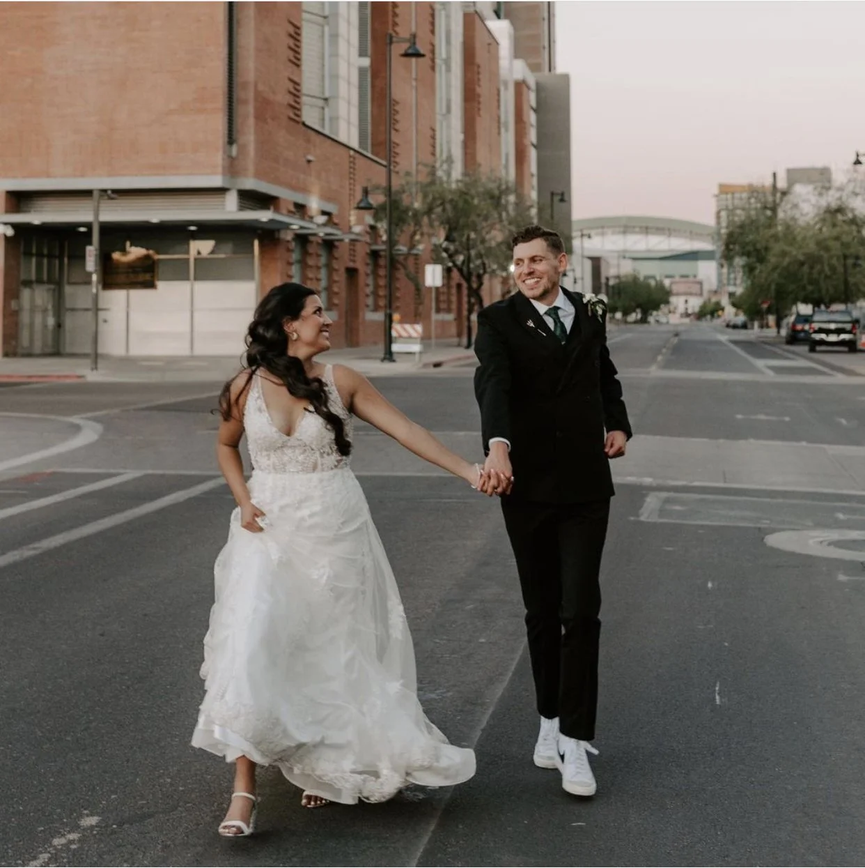 Bride and groom holding hands and walking on a city street, wearing wedding attire.
