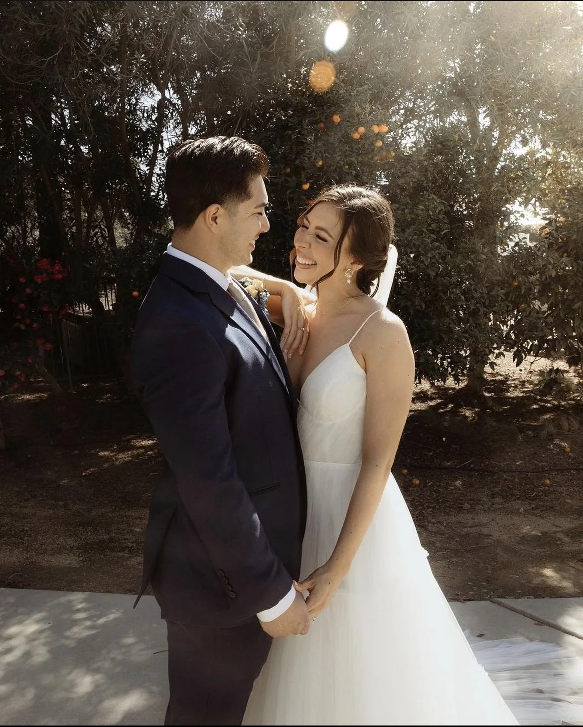 Bride and groom smiling and holding hands outdoors on their wedding day, standing against a backdrop of trees.