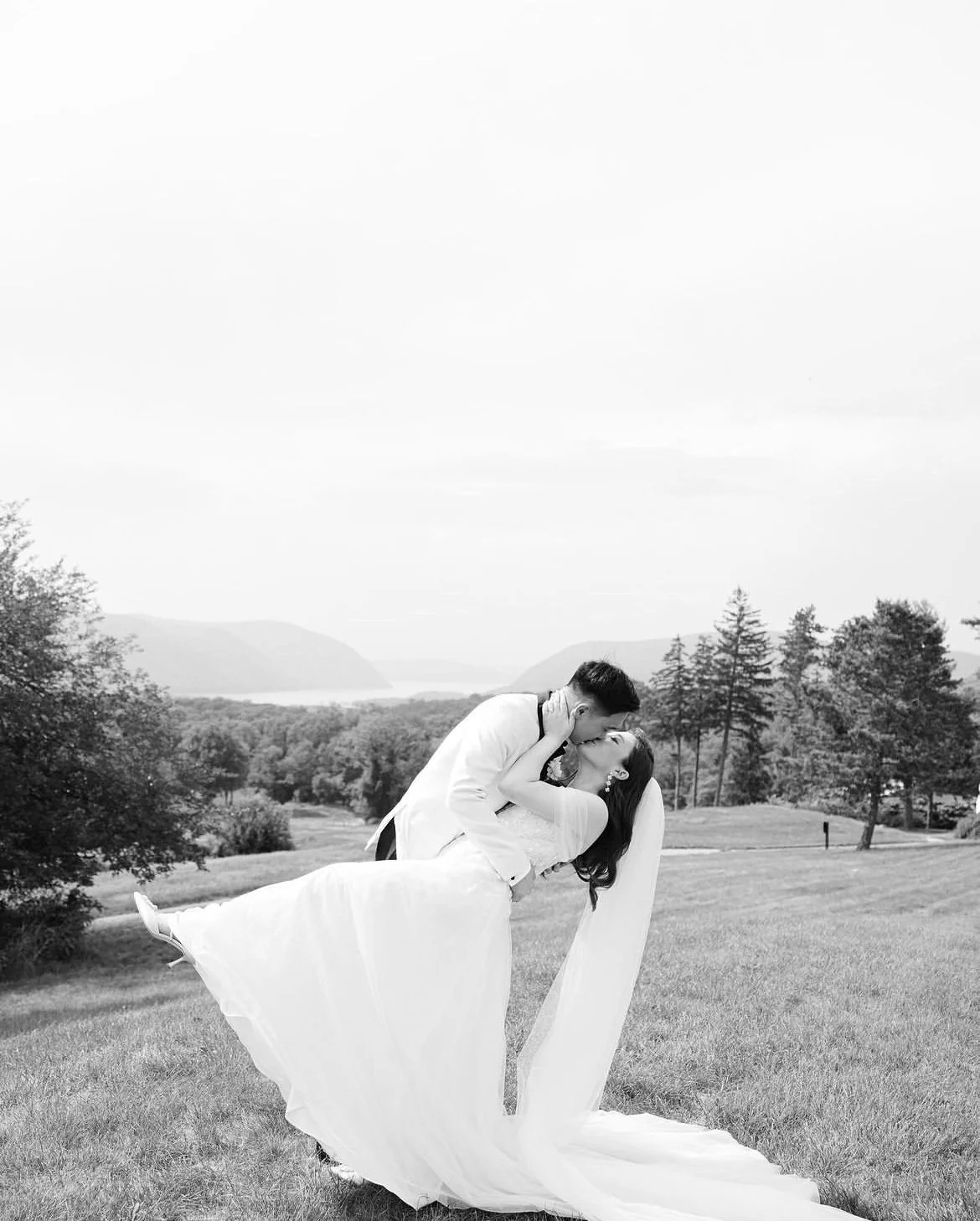 Bride and groom kissing in a scenic outdoor setting, with the groom dipping the bride, trees and mountains in the background.