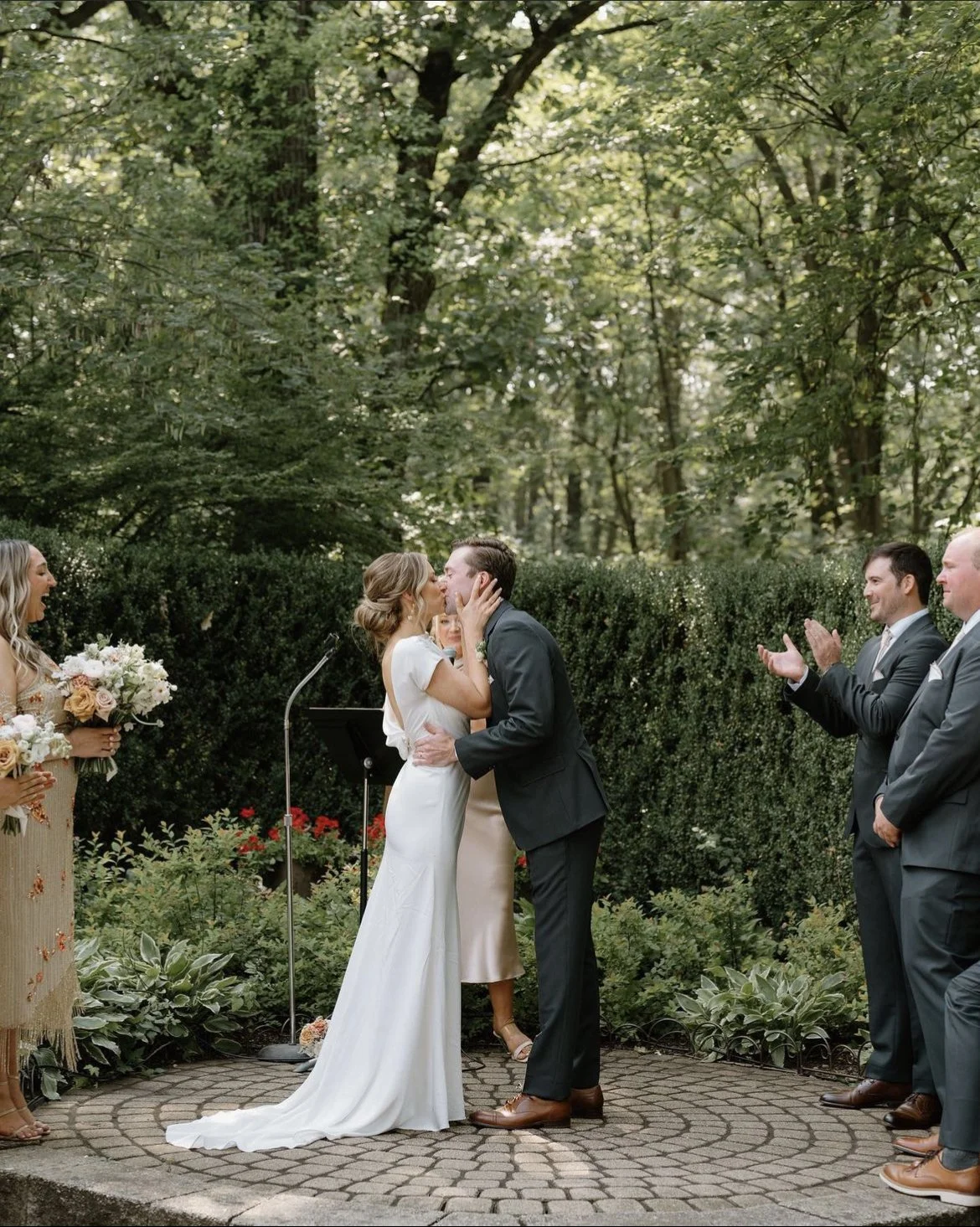 Bride and groom kissing during outdoor wedding ceremony with attendants and greenery.