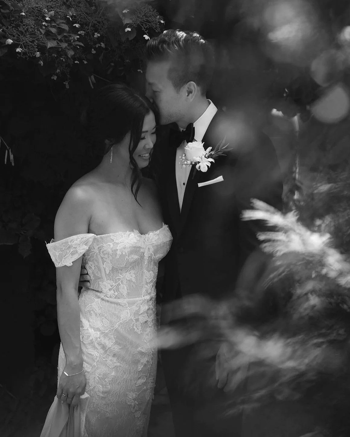 Black and white photo of a wedding couple; groom in a tuxedo kisses the bride in a lace dress.