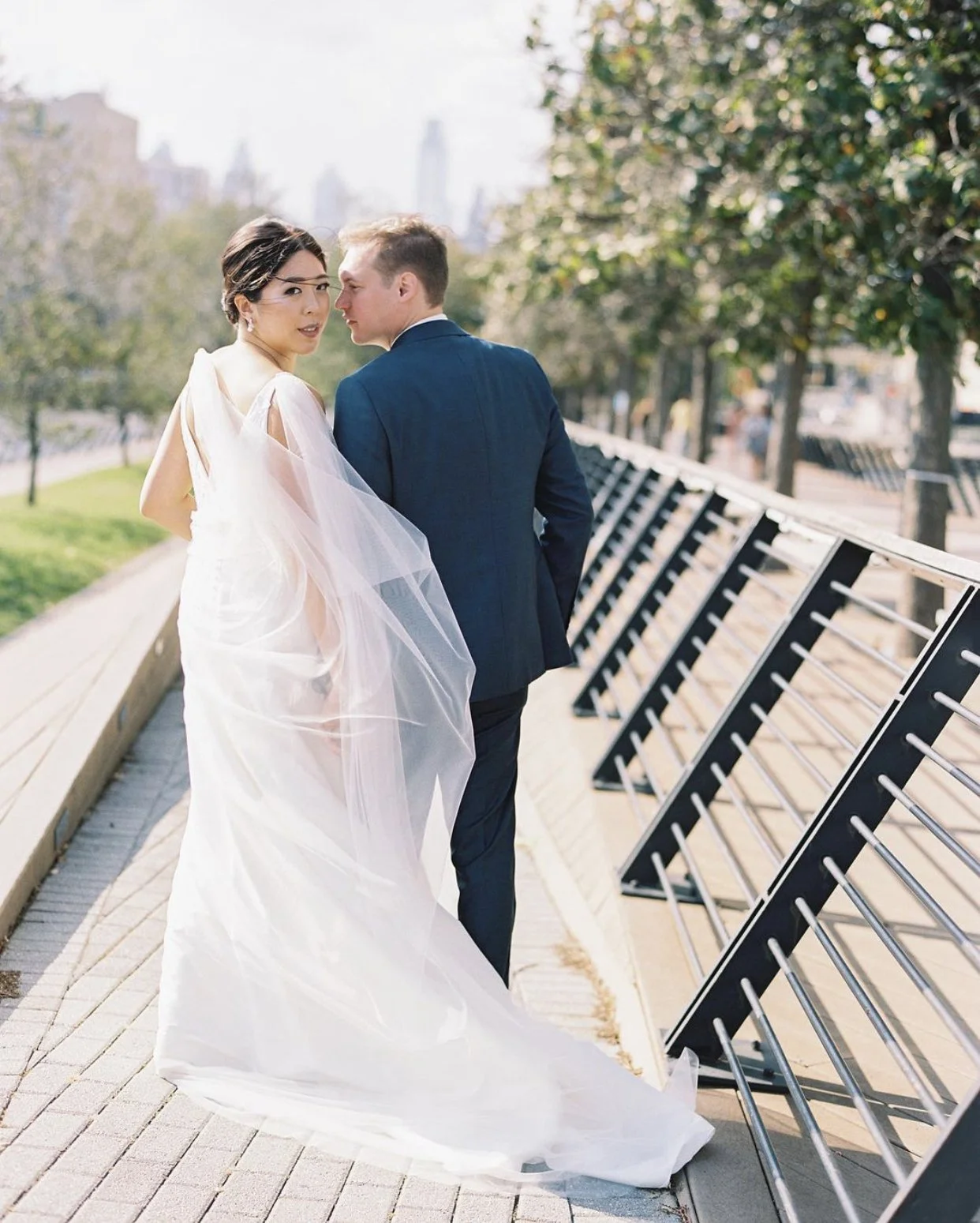 Bride and groom walking along a park path, with the bride in a white wedding dress and veil, and the groom in a dark suit, set against a cityscape background.