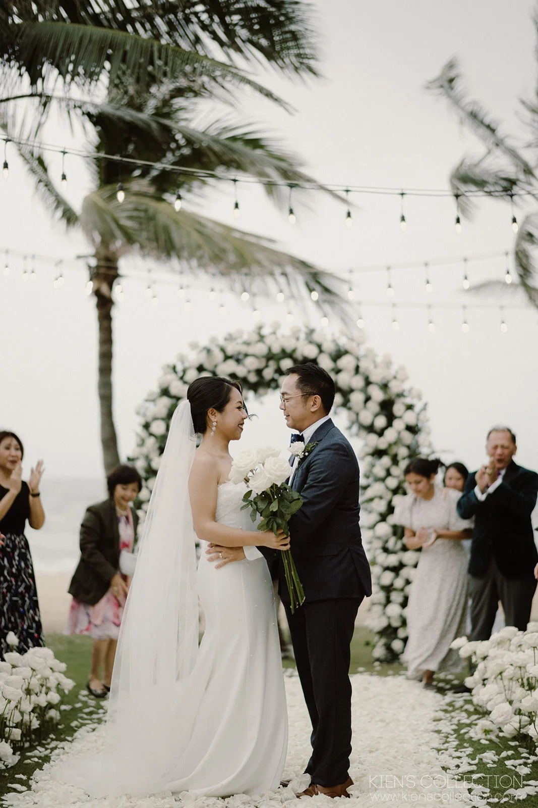 Bride and groom smiling at each other during an outdoor wedding ceremony by the beach, surrounded by white flowers and guests clapping.