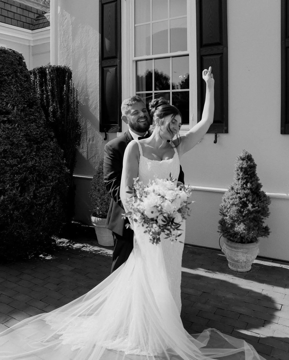 Bride and groom embracing outside, bride holding bouquet and raising hand, black and white photo.
