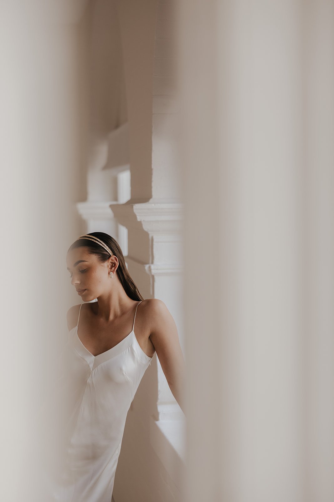 Woman in a white dress with a headband, standing in a softly lit, minimalist setting with architectural details.