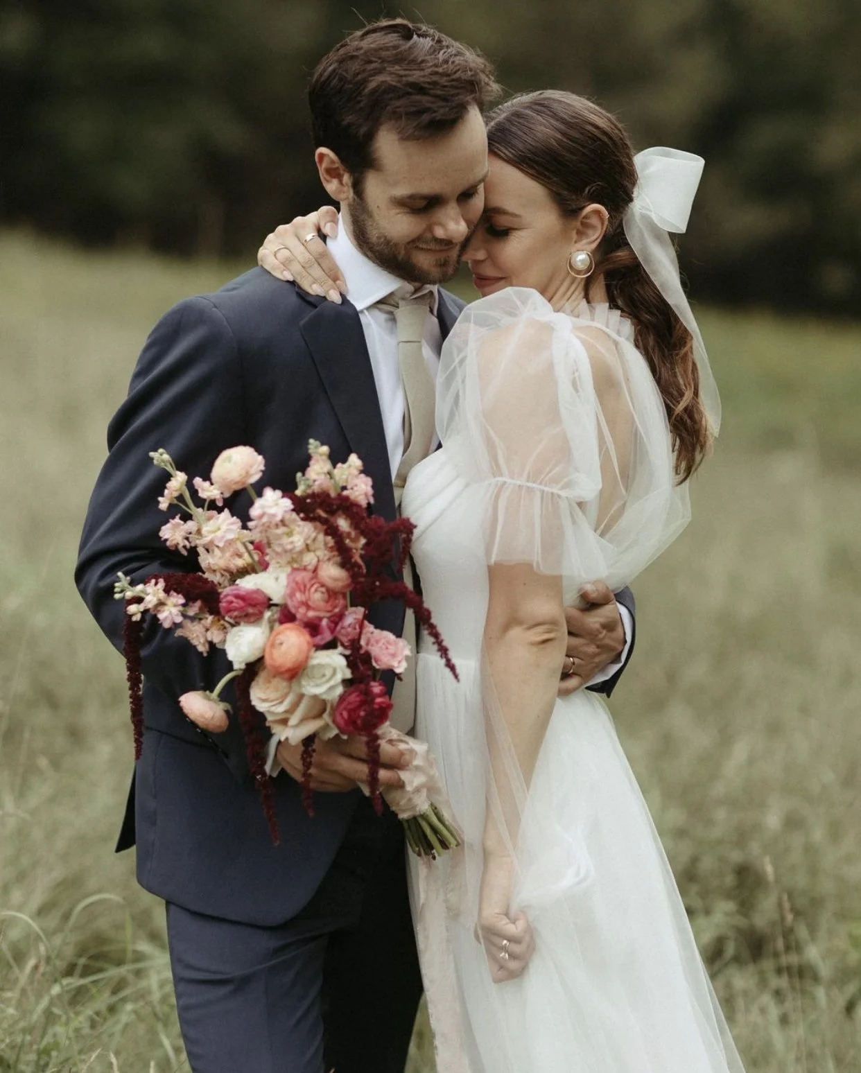 A couple embracing at their wedding, the groom in a blue suit and the bride in a white dress holding a bouquet of colorful flowers, in an outdoor setting.