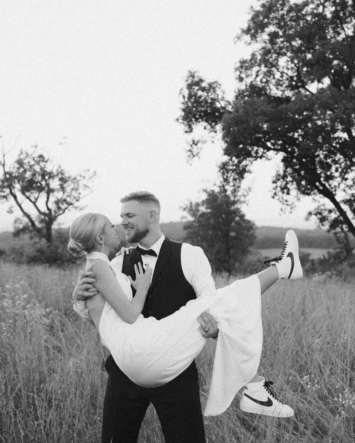 A man in formal attire holds a woman wearing a white dress and sneakers, standing in a field with trees in the background. Black and white image.