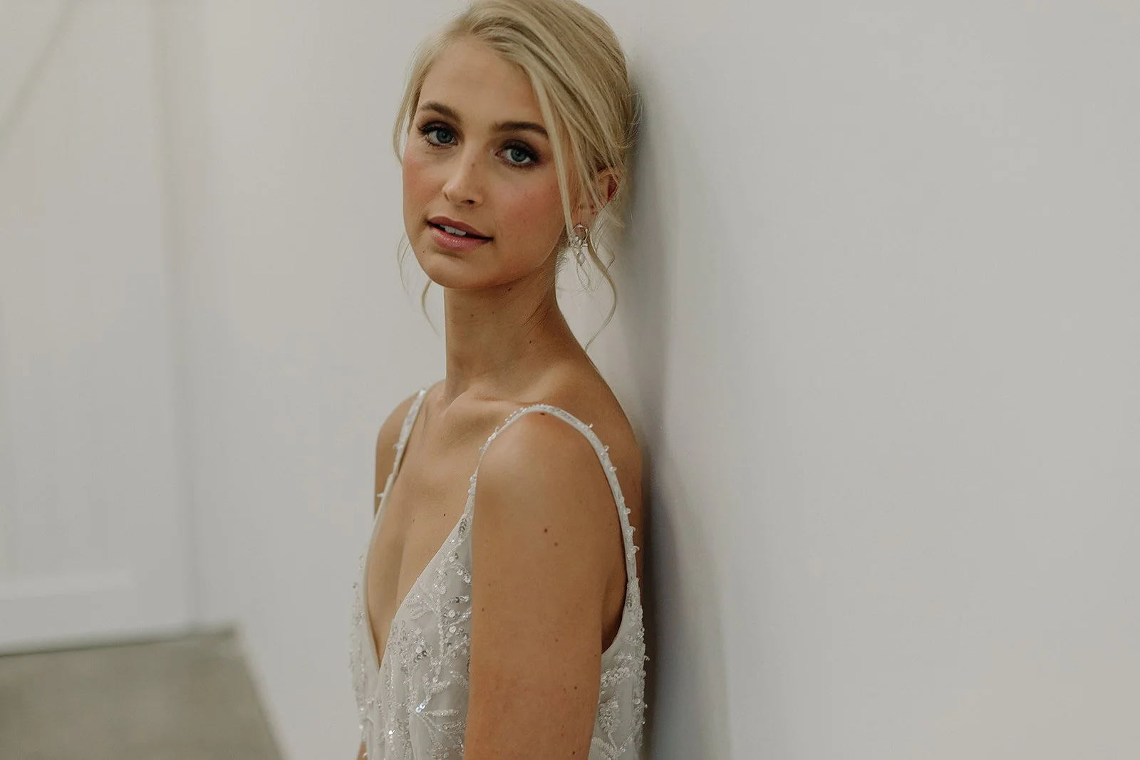 A woman in a white dress leaning against a wall, looking at the camera. She has blonde hair styled in an updo.