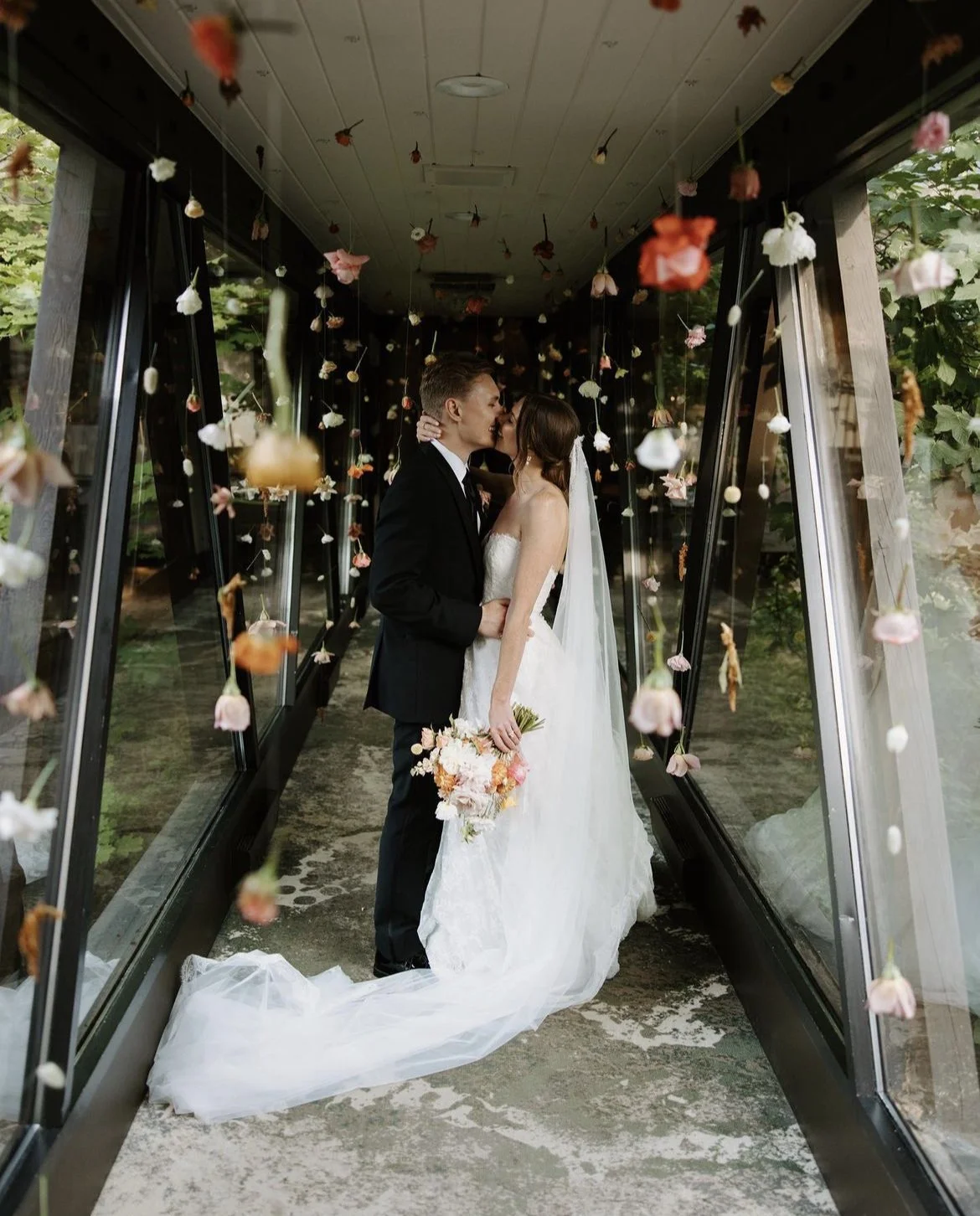 Bride and groom kissing in a corridor with suspended flowers.