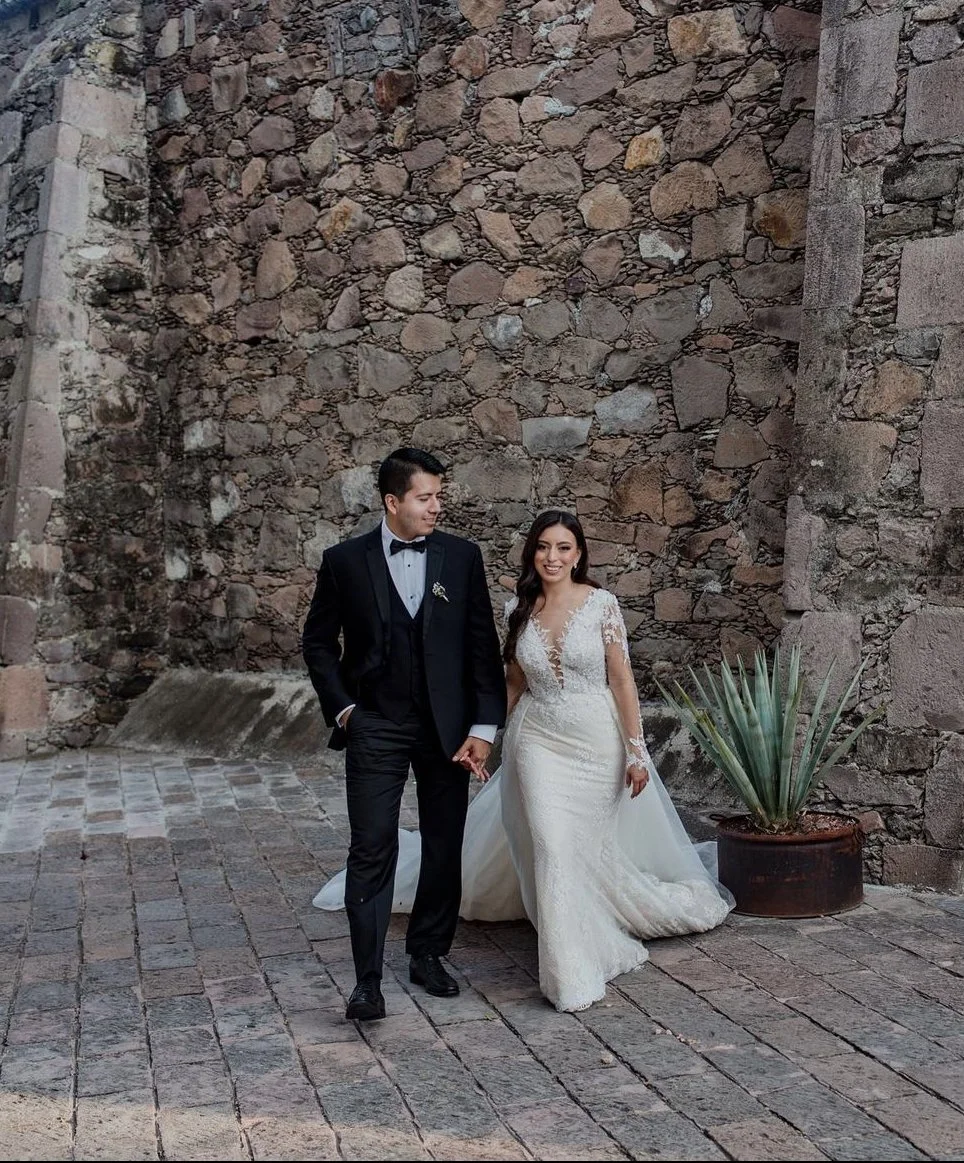 Bride and groom walking next to a rustic stone wall, with a potted plant nearby.