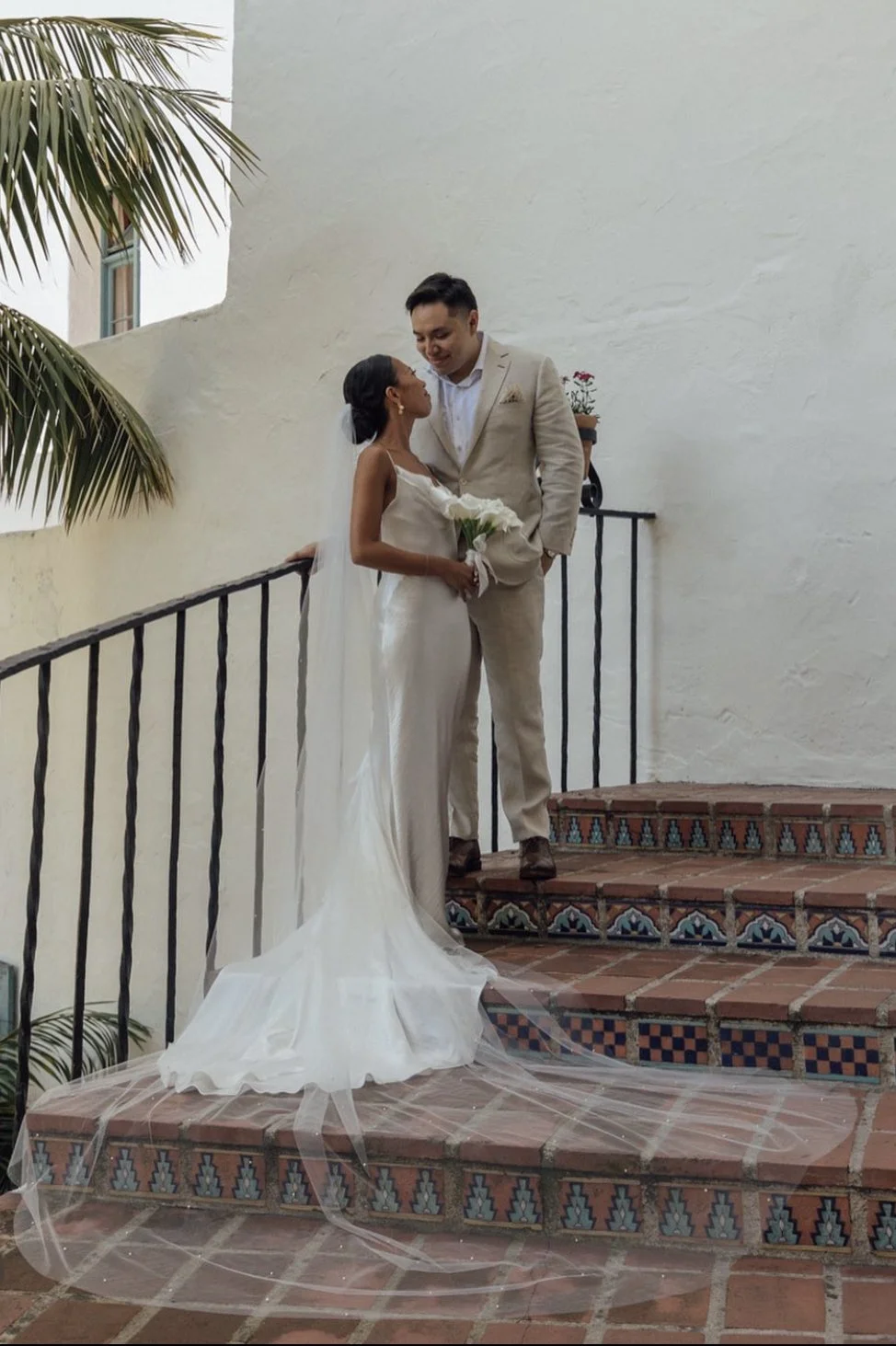 Bride and groom on tiled staircase, bride holding bouquet, groom in light suit, wedding scene with tropical plant and white wall.