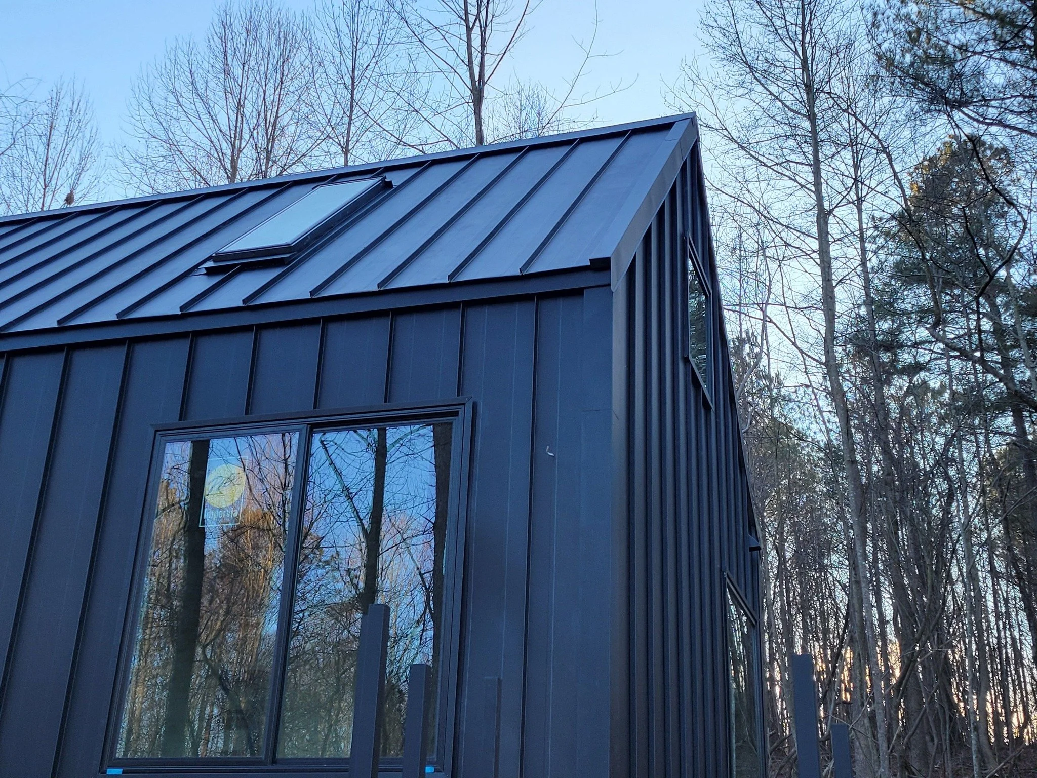 A modern black metal house with large windows and a skylight, surrounded by leafless trees during twilight.