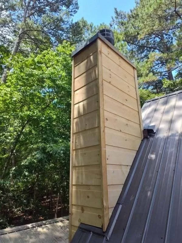 Wooden chimney on a metal roof surrounded by trees.