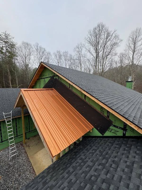 A house under construction with different roofing styles, including black shingles, copper-colored metal, and black flashing, with a ladder leaning against the side.