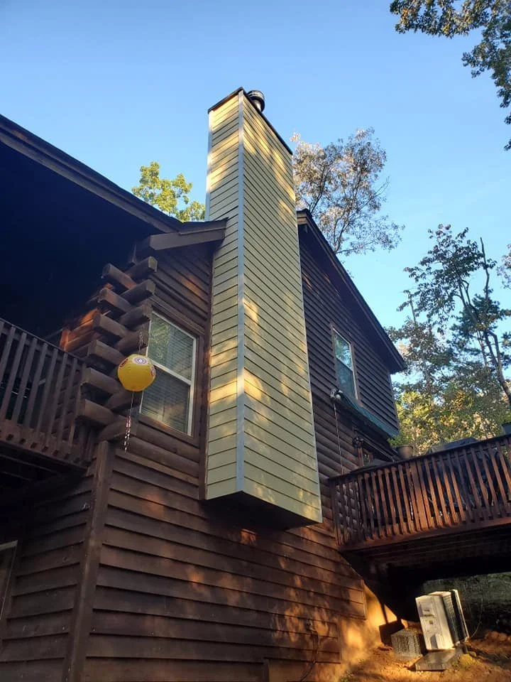 View of a house with brown wood siding and a tall, light-colored exterior chimney or vent pipe extending above the roof, with trees and blue sky in the background.