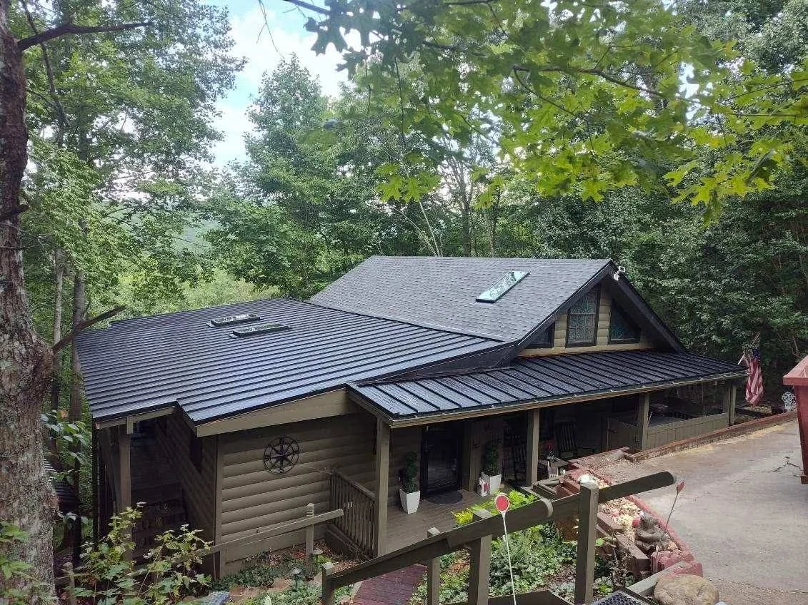 A house with a sloped roof surrounded by green trees, with a small porch and seating area in front.