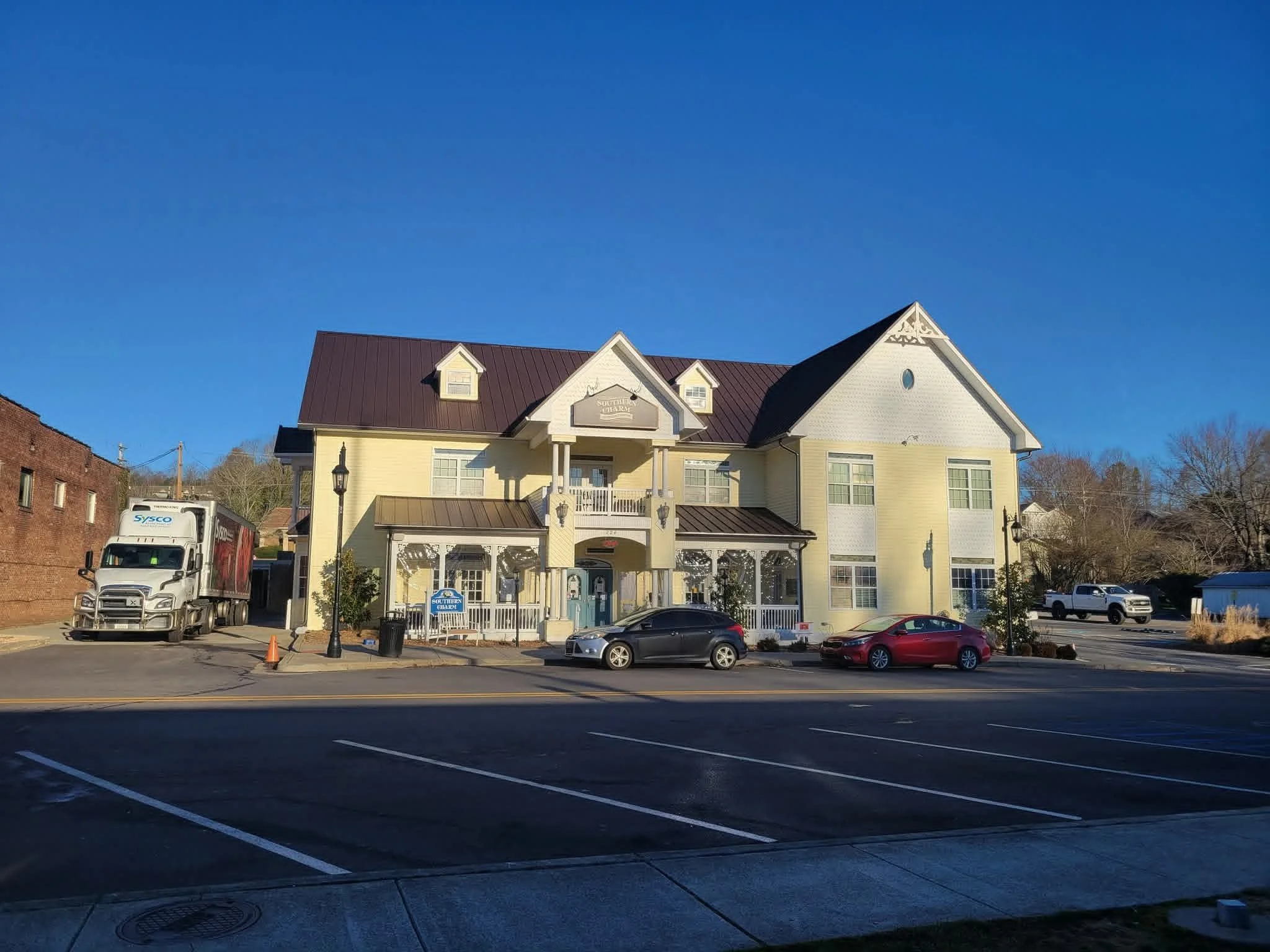 A large yellow Victorian-style building with white trim and a dark brown roof, situated behind a parking lot with several cars and a semi-truck, under a clear blue sky.