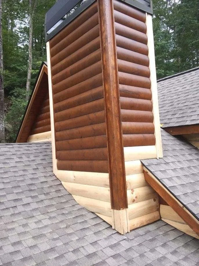 Close-up of a log cabin-style chimney on a house with asphalt shingle roof, surrounded by trees.