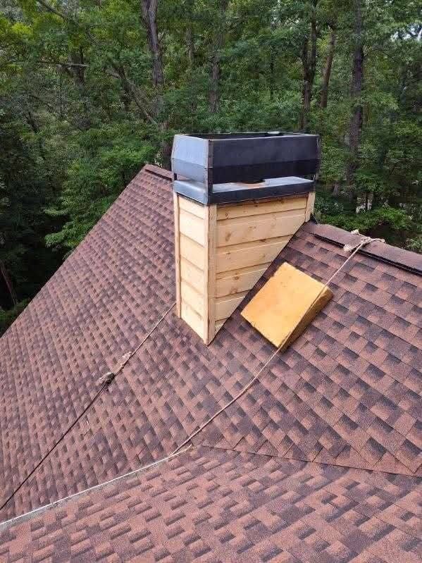 Chimney on a roof with a black cap, made of wood panels, with trees in the background.