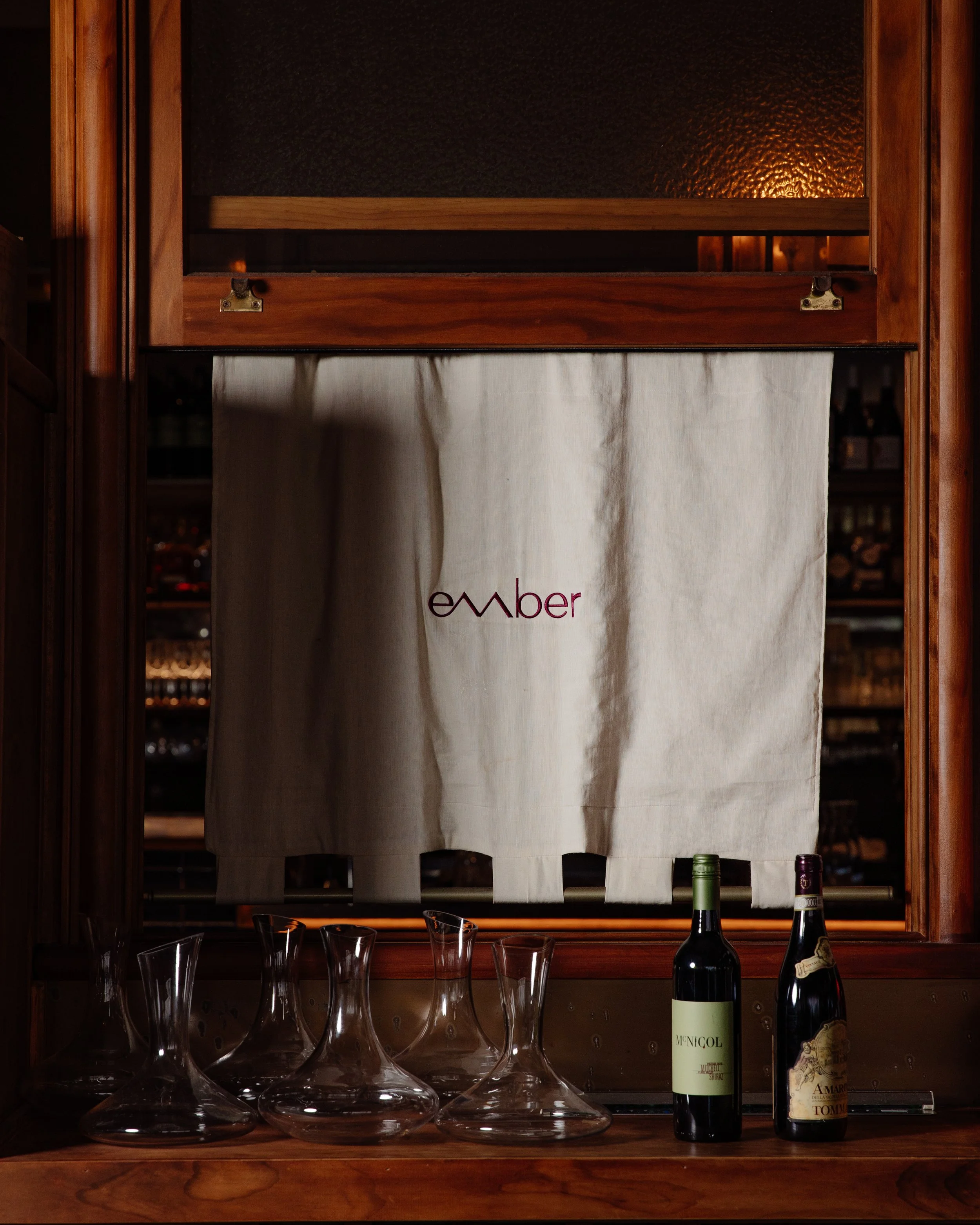 A wooden bar shelf with empty decanters, two bottles of wine, and a white curtain with red text partially visible behind the shelf.