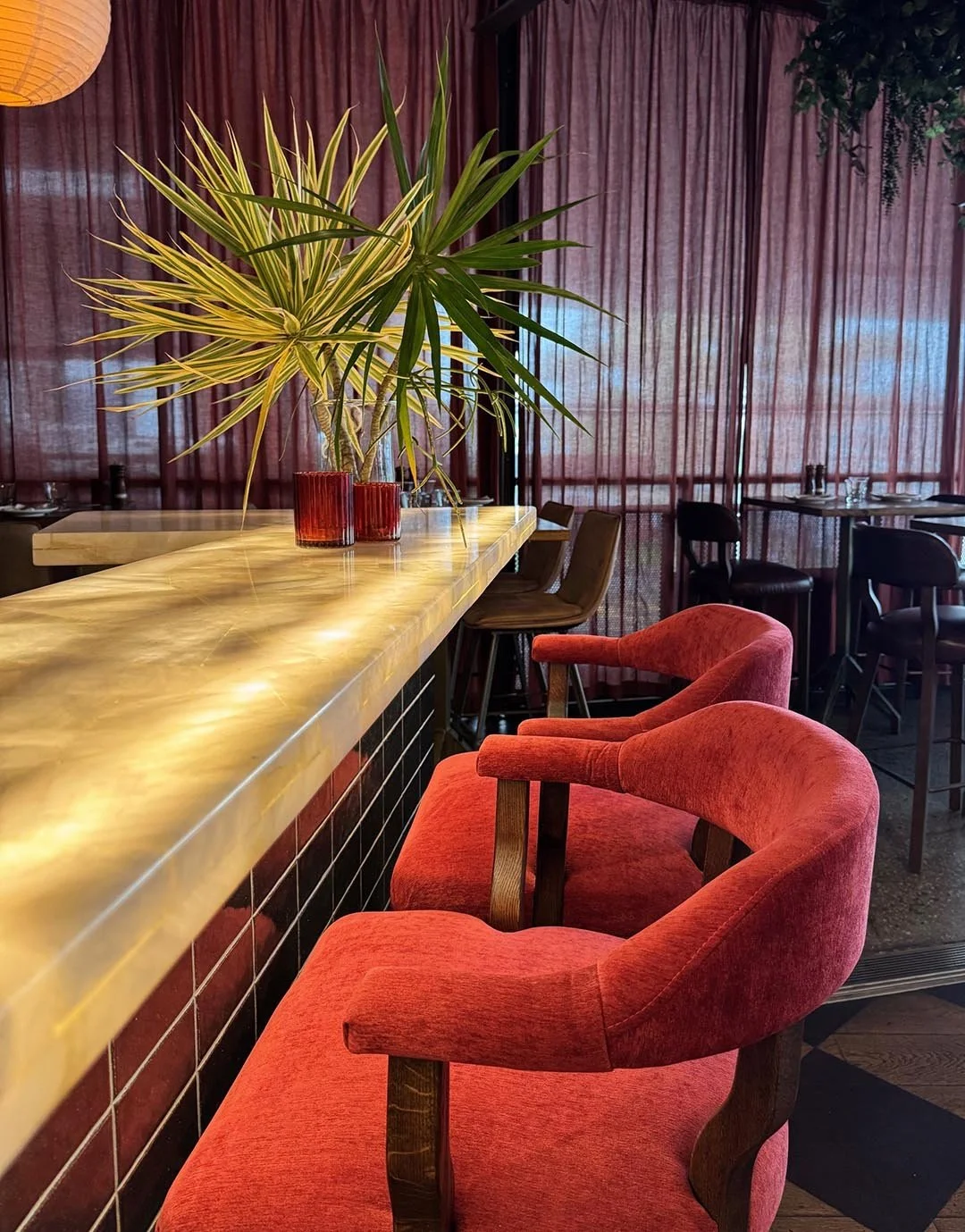 Interior of a restaurant with red velvet chairs, a marble-top bar, and a large potted plant on the counter.