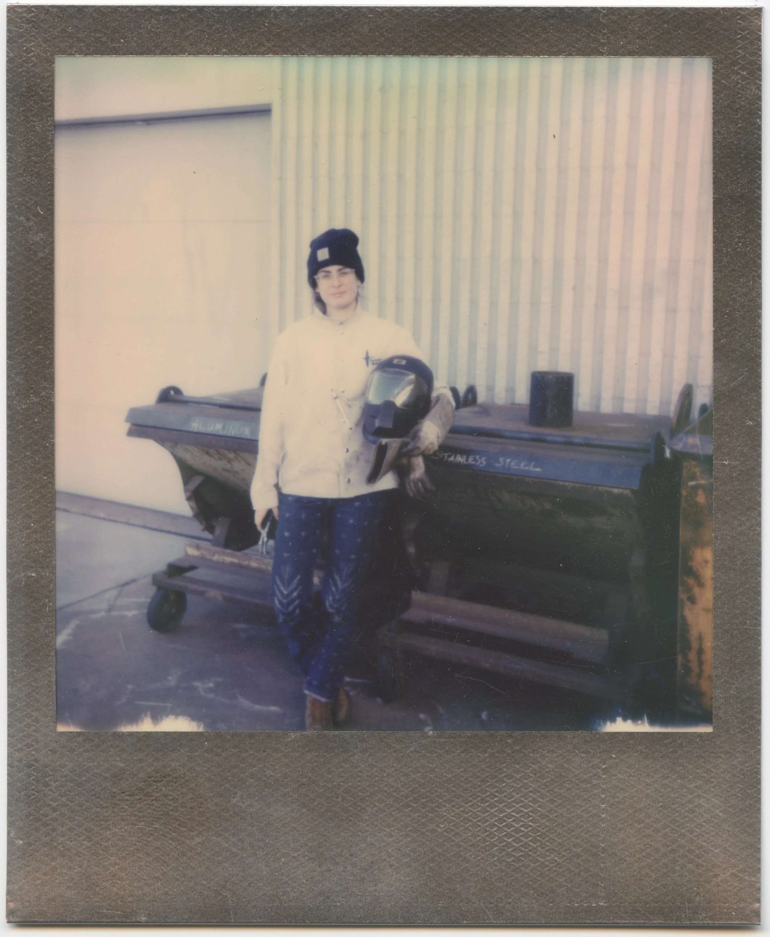 Self portrait by the recycling bins. Alex Matzke is photographed with a polaroid camera. Alex went to industrial welding school and learned alongside pipefitters.