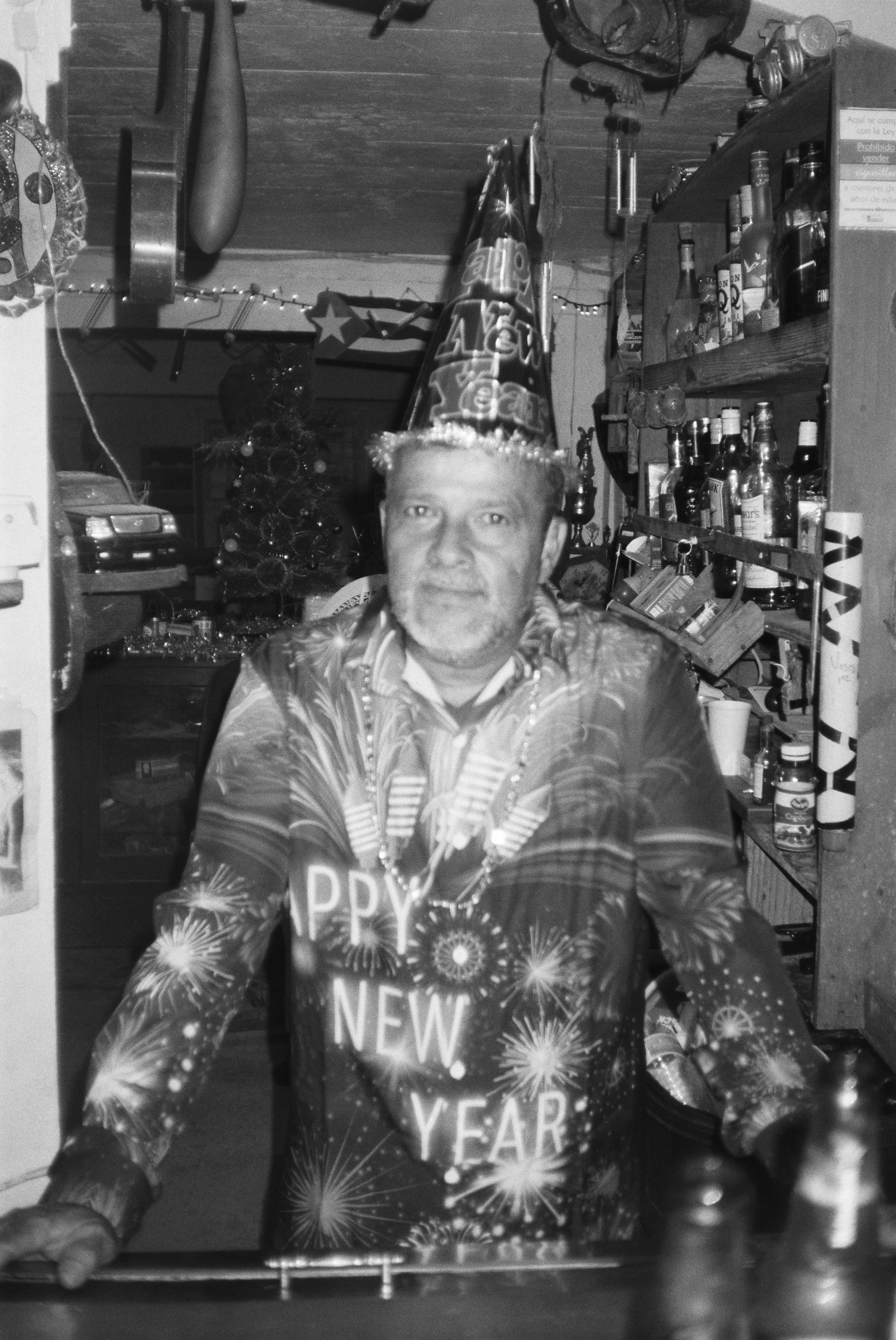 Sammie stands behind the bar, surrounded by decorations and trinkets, and a full liquor cabinet. He is festively dressed for new years, and sports and hat and themed shirt.