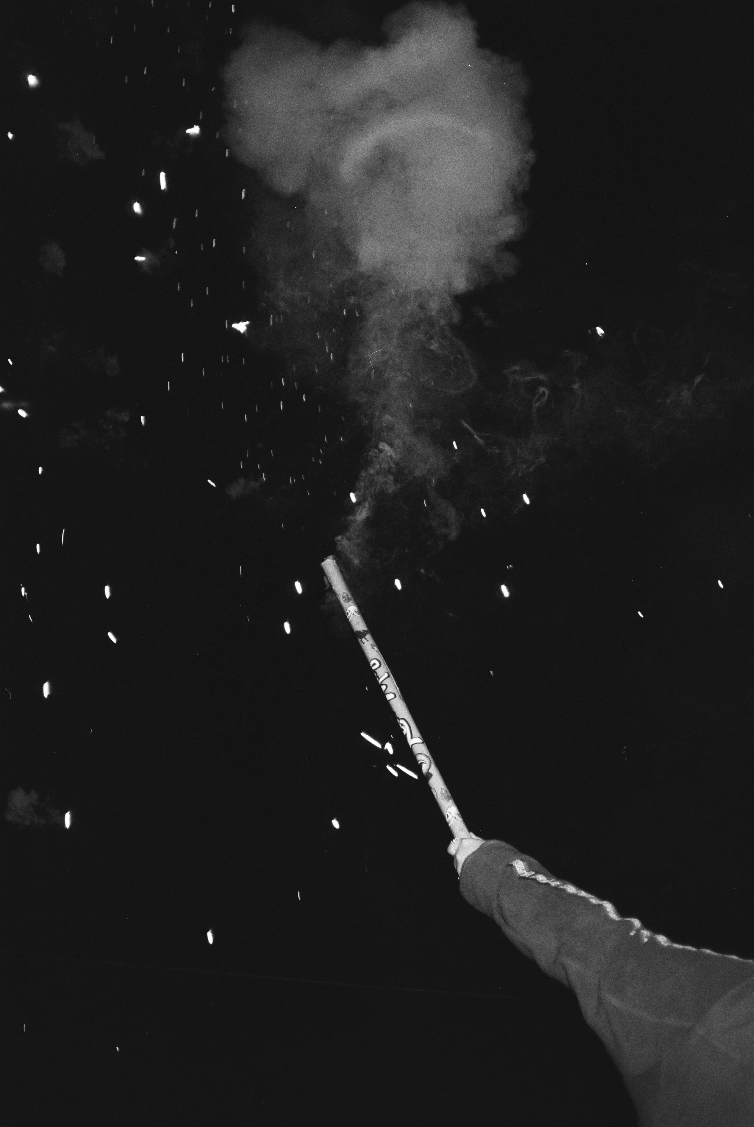 someone holds a Roman candle style firework in their hand in this black and white 35mm image. The firework has just gone off, and there are still sparks and smoke visible in the image.